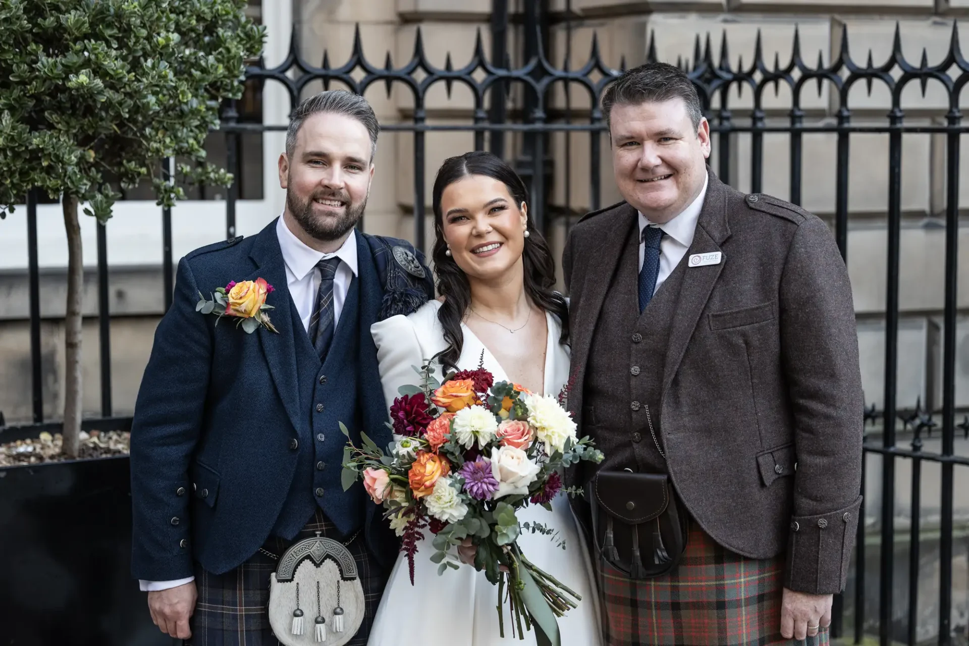Three people dressed in formal attire, including two men in kilts and a woman holding a bouquet, smiling together at a wedding event.