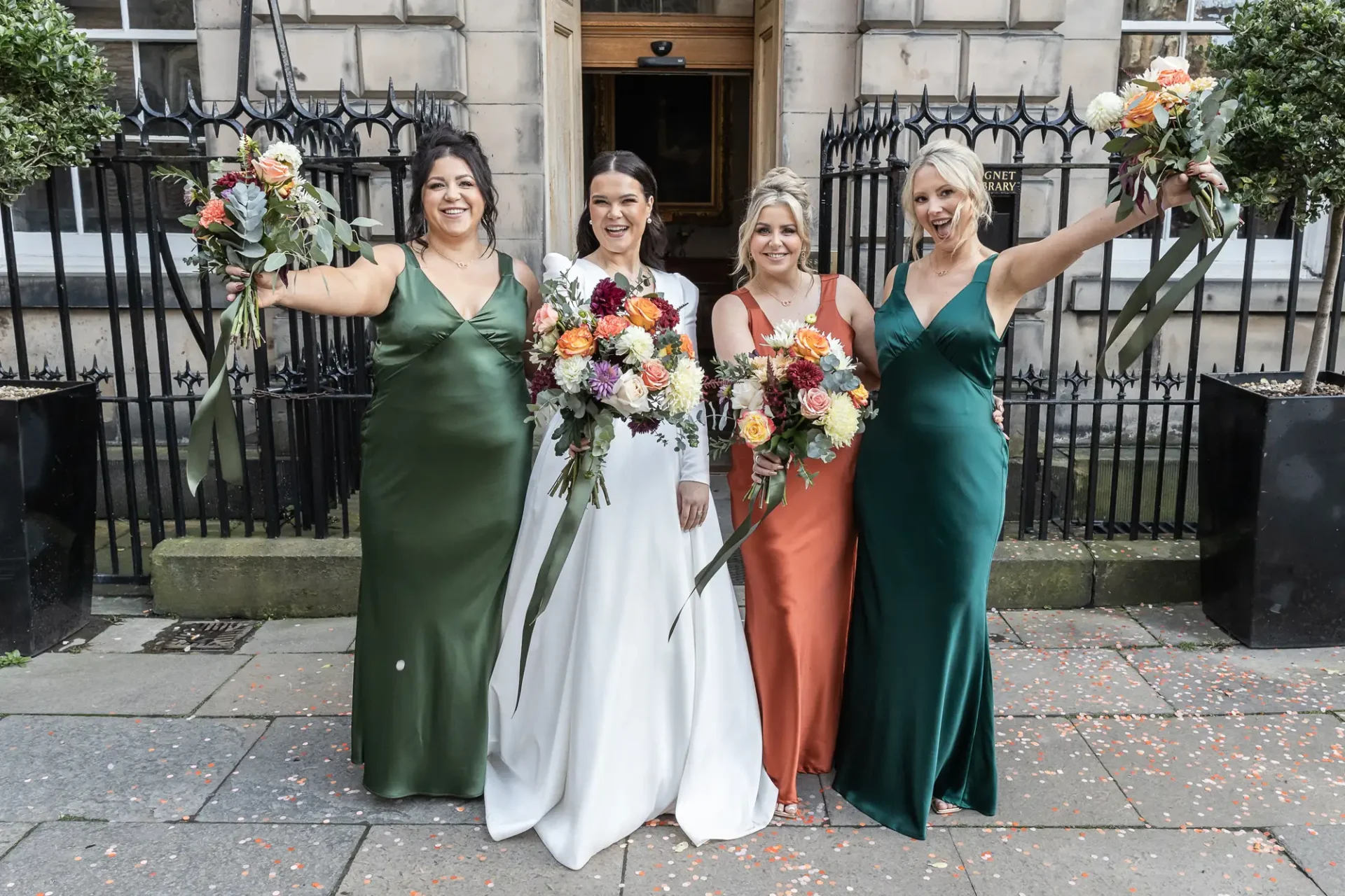 Four women in formal dresses, one in white and three in green, hold large bouquets and pose smiling on a city street.