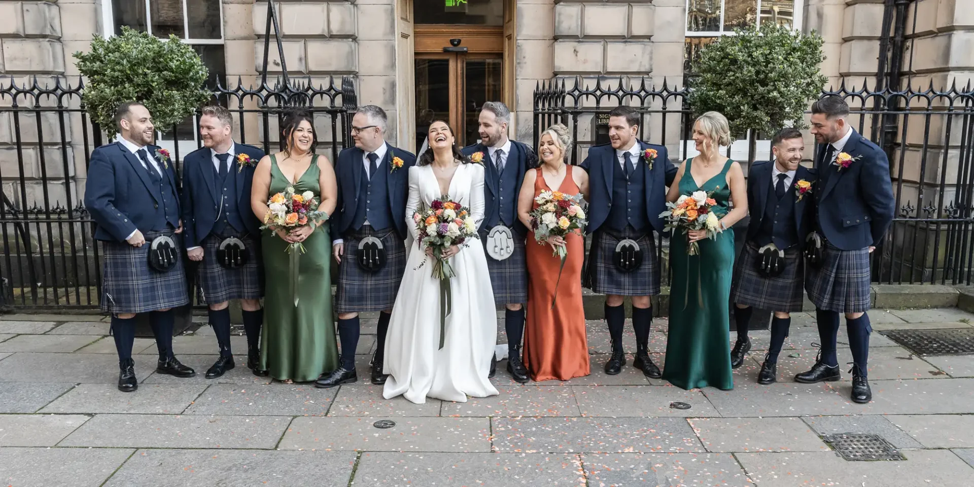 Wedding group laughing together on city sidewalk, featuring several men in kilts and women in dresses, with bouquets.