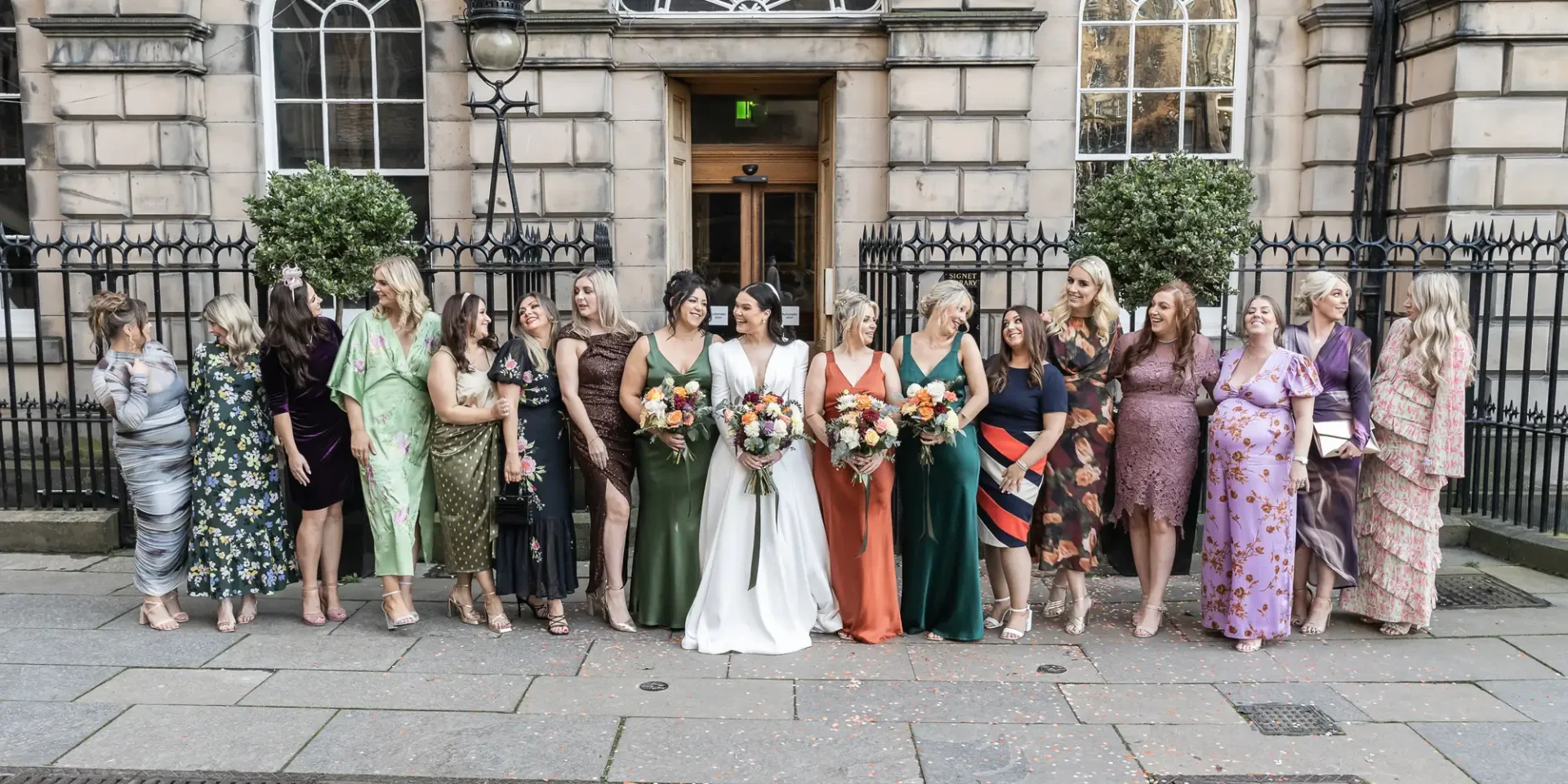A bride in a white dress flanked by bridesmaids in colorful dresses and guests, holding bouquets, posing outside an elegant building.