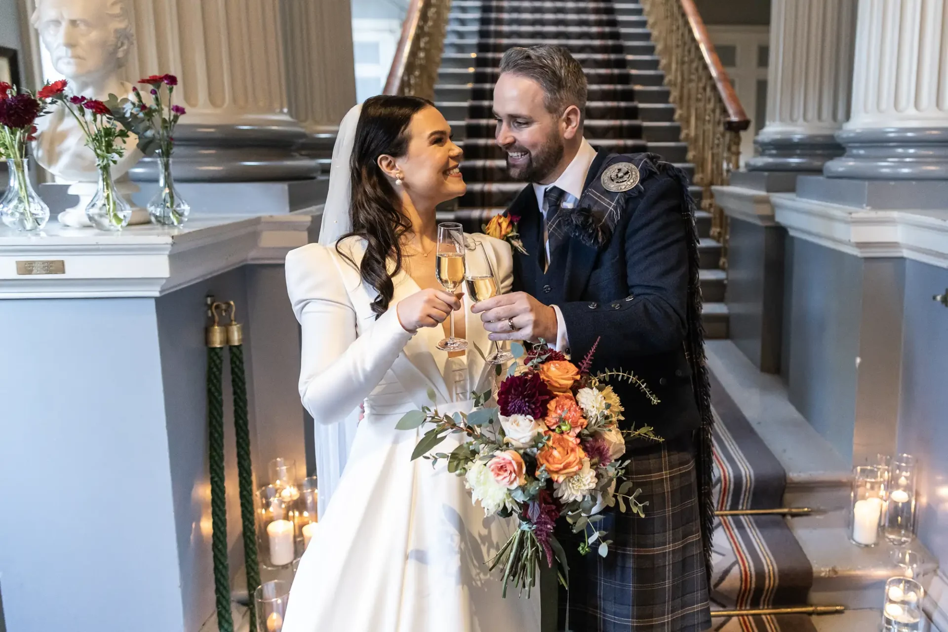 A bride and groom in formal attire, toasting with champagne on a staircase decorated with candles and flowers.