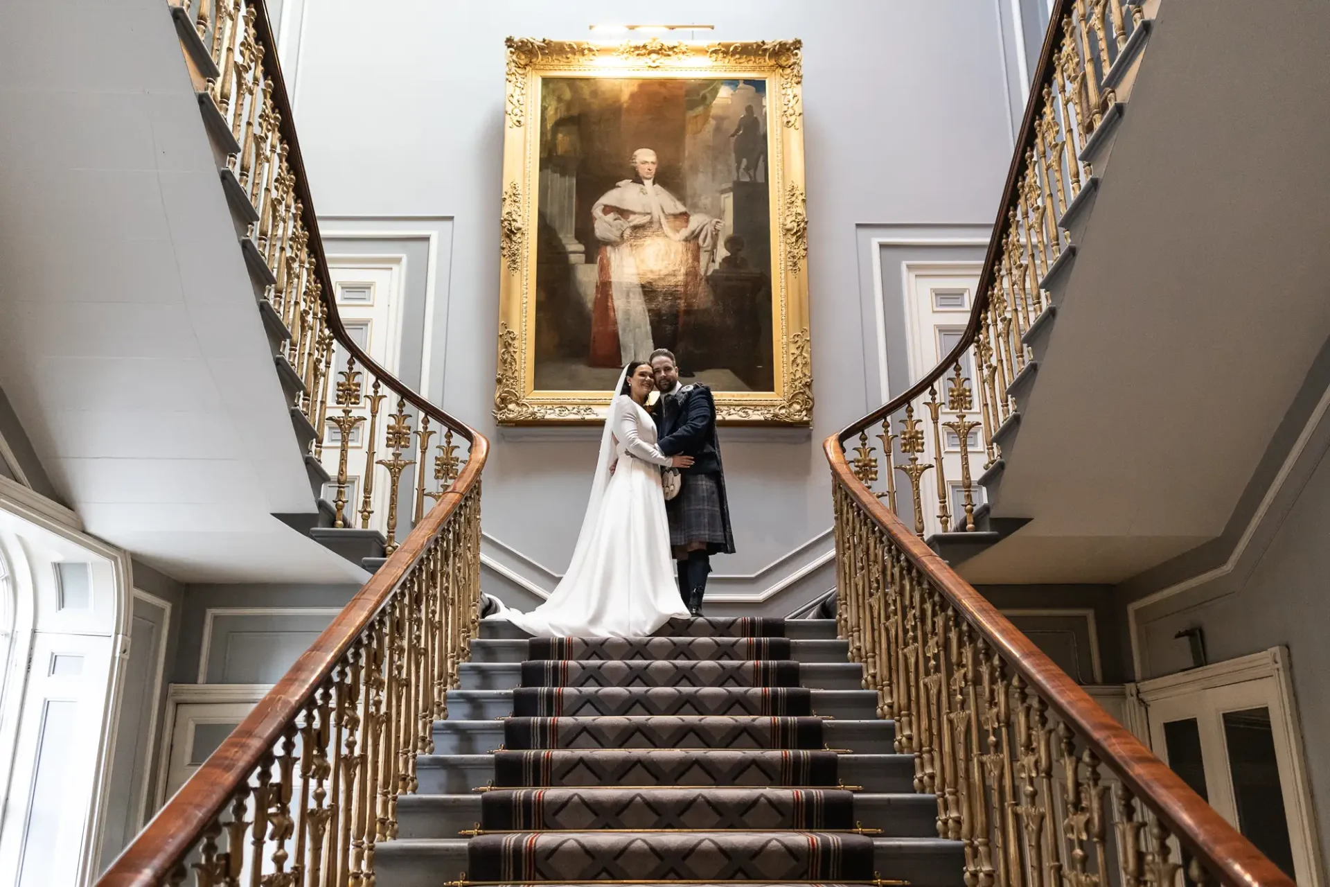 A newlywed couple embraces on an ornate staircase under a large portrait in a grand hall.