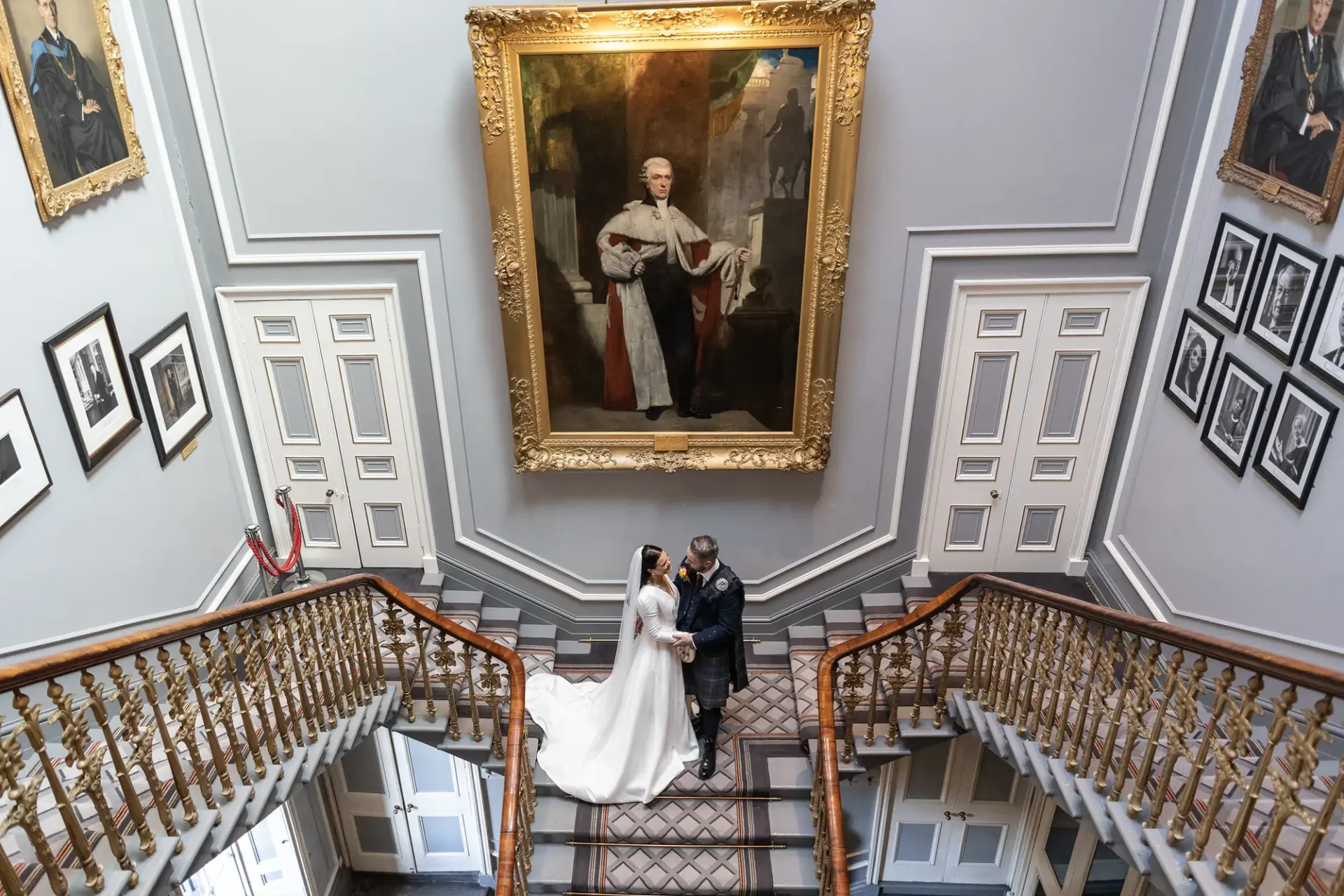 A bride and groom holding hands stand at the base of a grand staircase, flanked by classical paintings in an elegant hall.