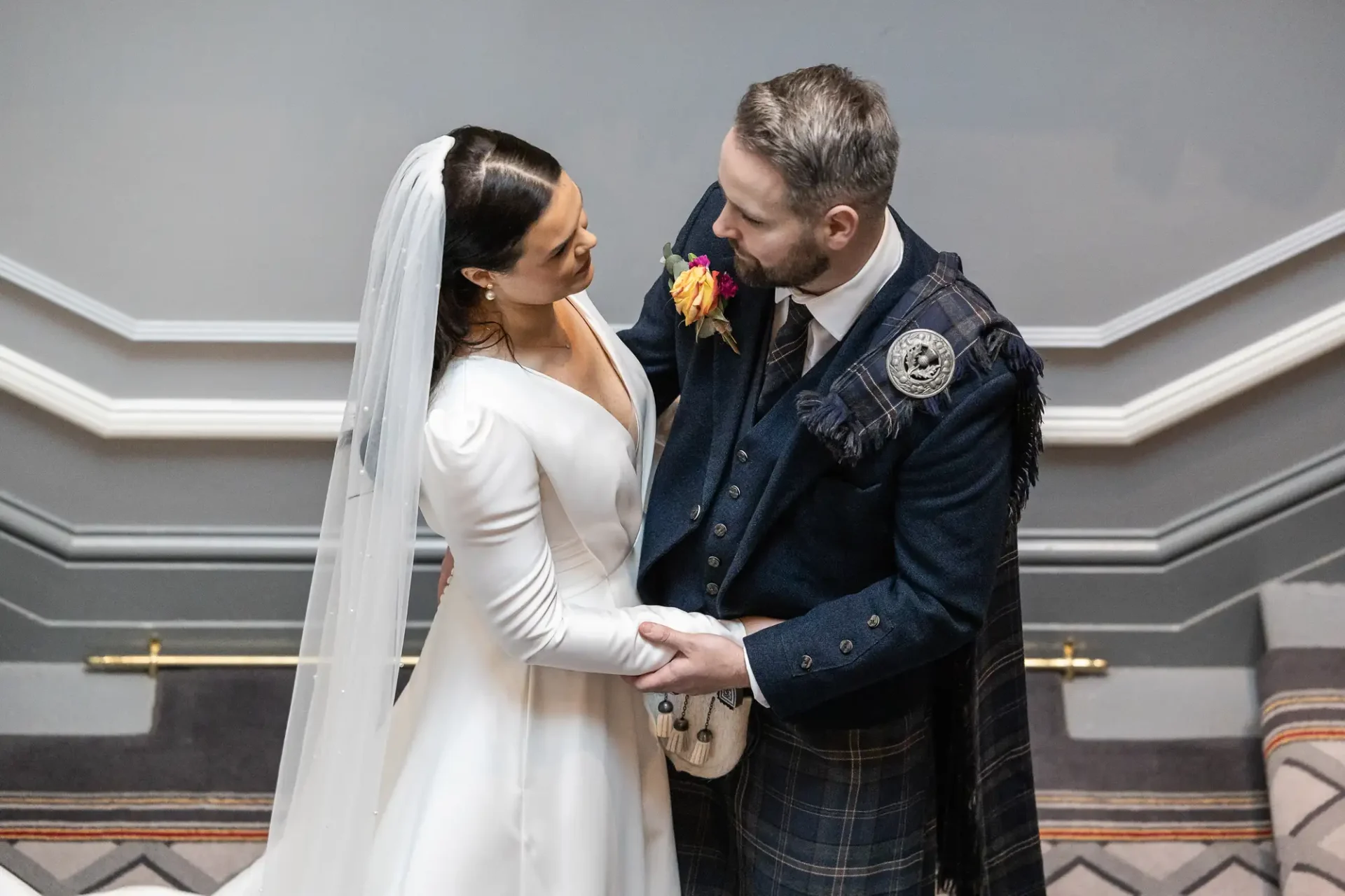 Bride in a white dress and groom in a kilt sharing a tender moment on a staircase, touching foreheads lovingly.