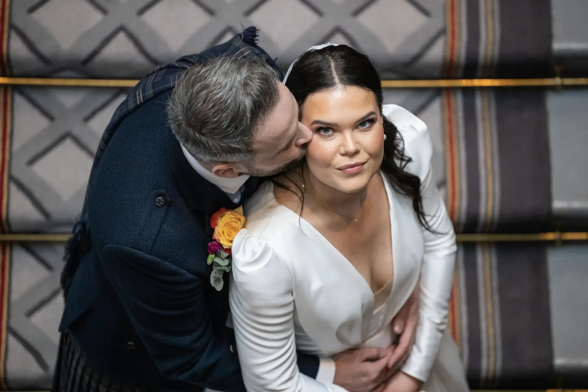 A groom in a dark suit kisses the cheek of a bride in a white dress. they are sitting on a patterned couch with a warm embrace.