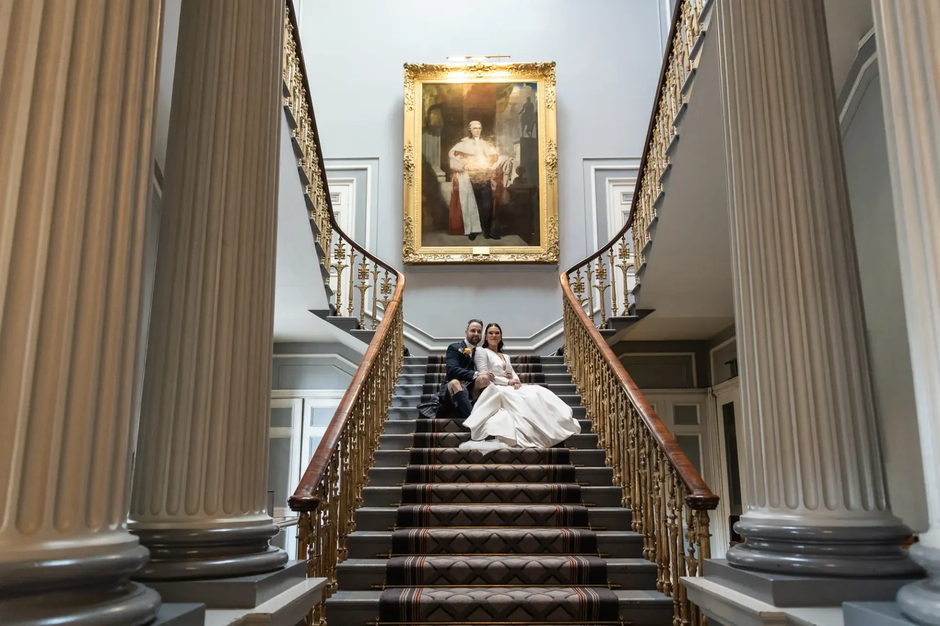 A bride and groom pose on an elegant staircase underneath a large portrait in a grand hall.