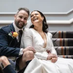 A joyful couple in wedding attire sitting on a staircase, the man in a kilt and the woman in a white dress, both laughing.