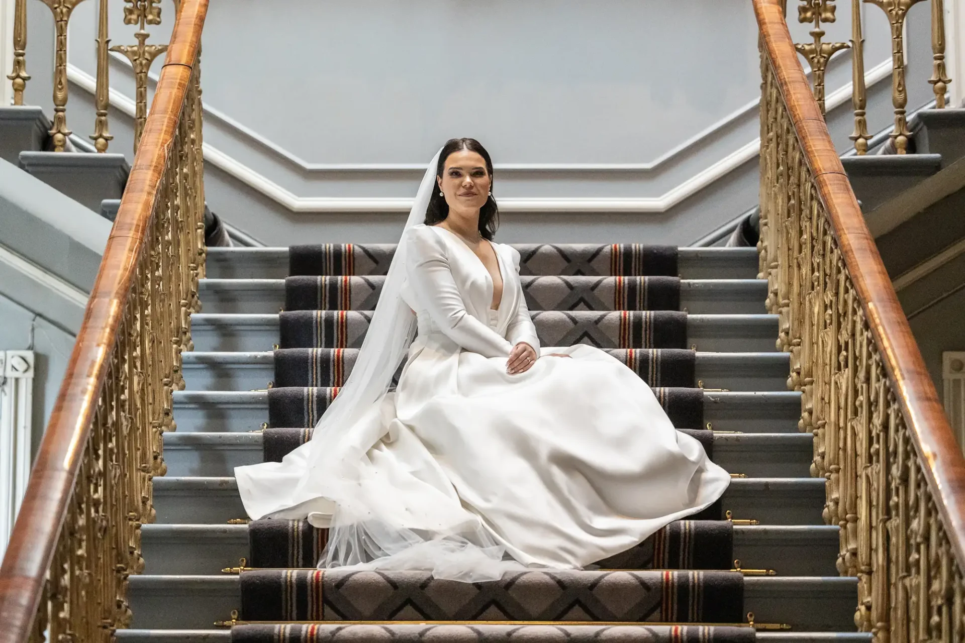 A bride in a white dress smiles while sitting on a staircase with ornate railings in an elegant interior.
