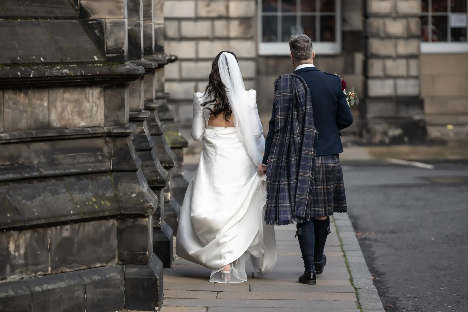 A bride in a white gown and a groom in a kilt walk together along a city street, viewed from behind.