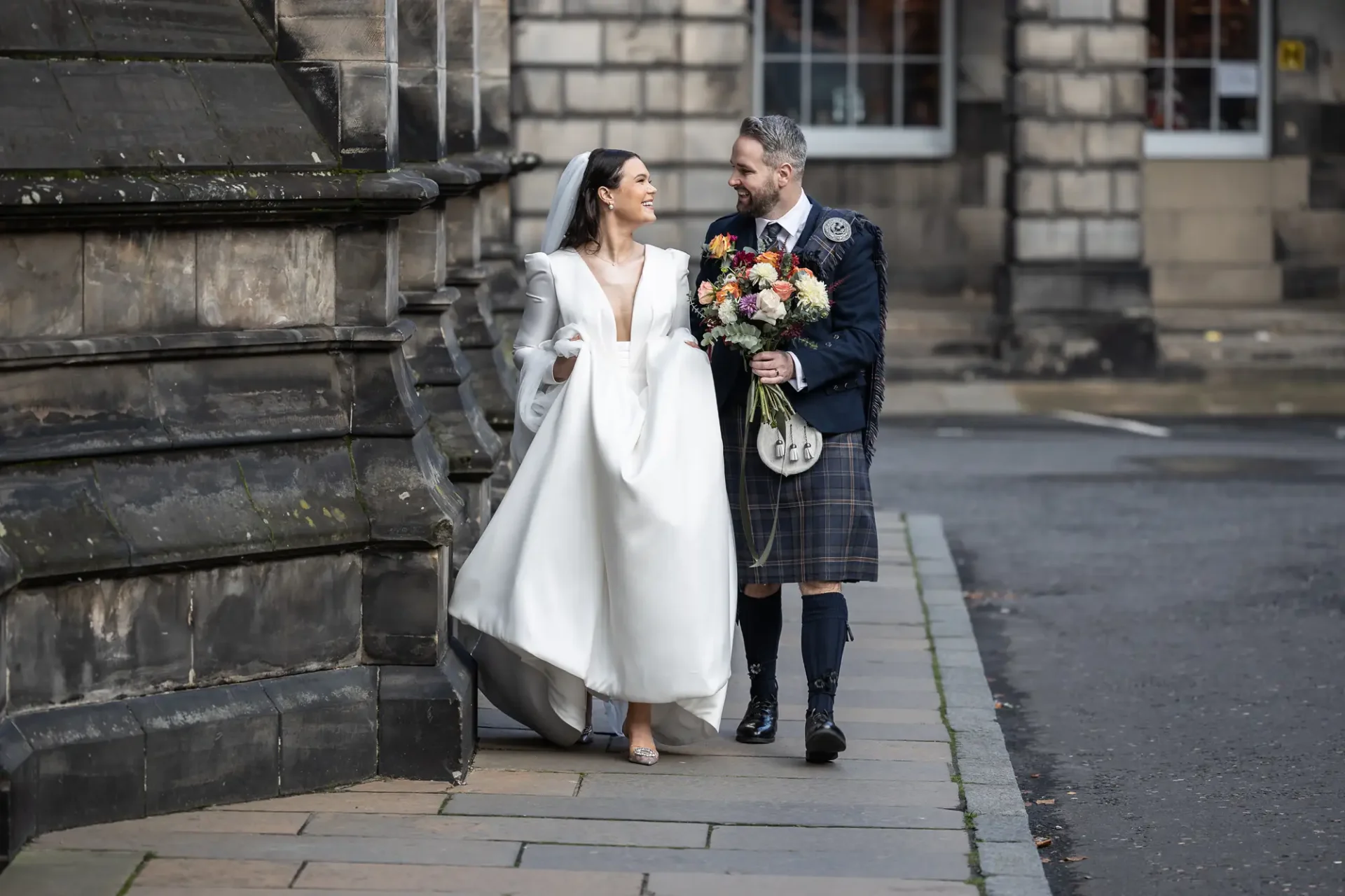 A bride in a white dress and a groom in a kilt, holding hands and walking along a city street, smiling at each other.