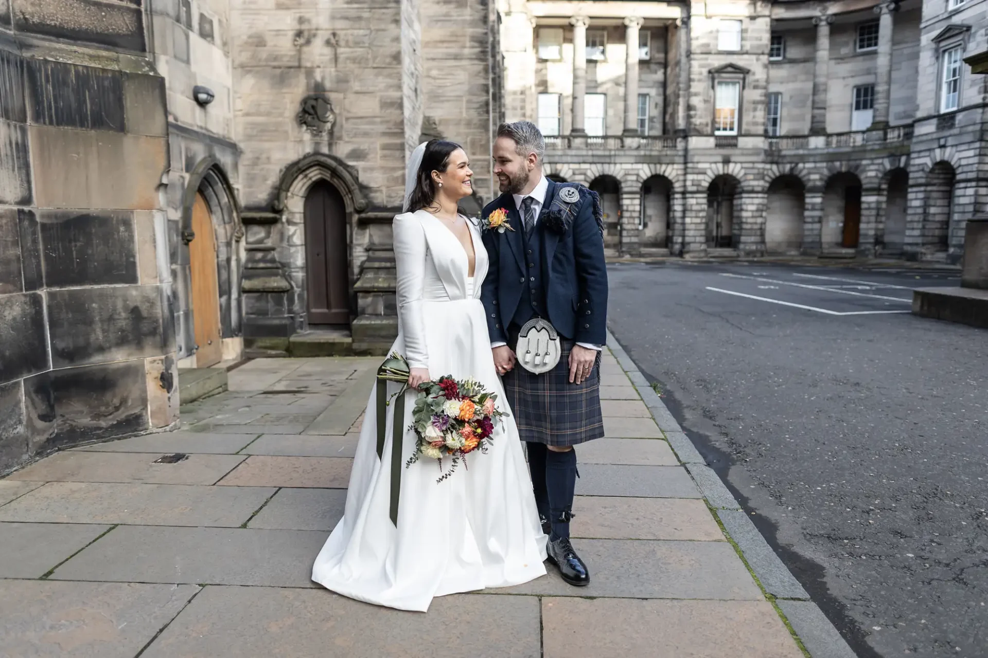 A bride in a white dress and a groom in a kilt smiling at each other, holding hands in a courtyard.