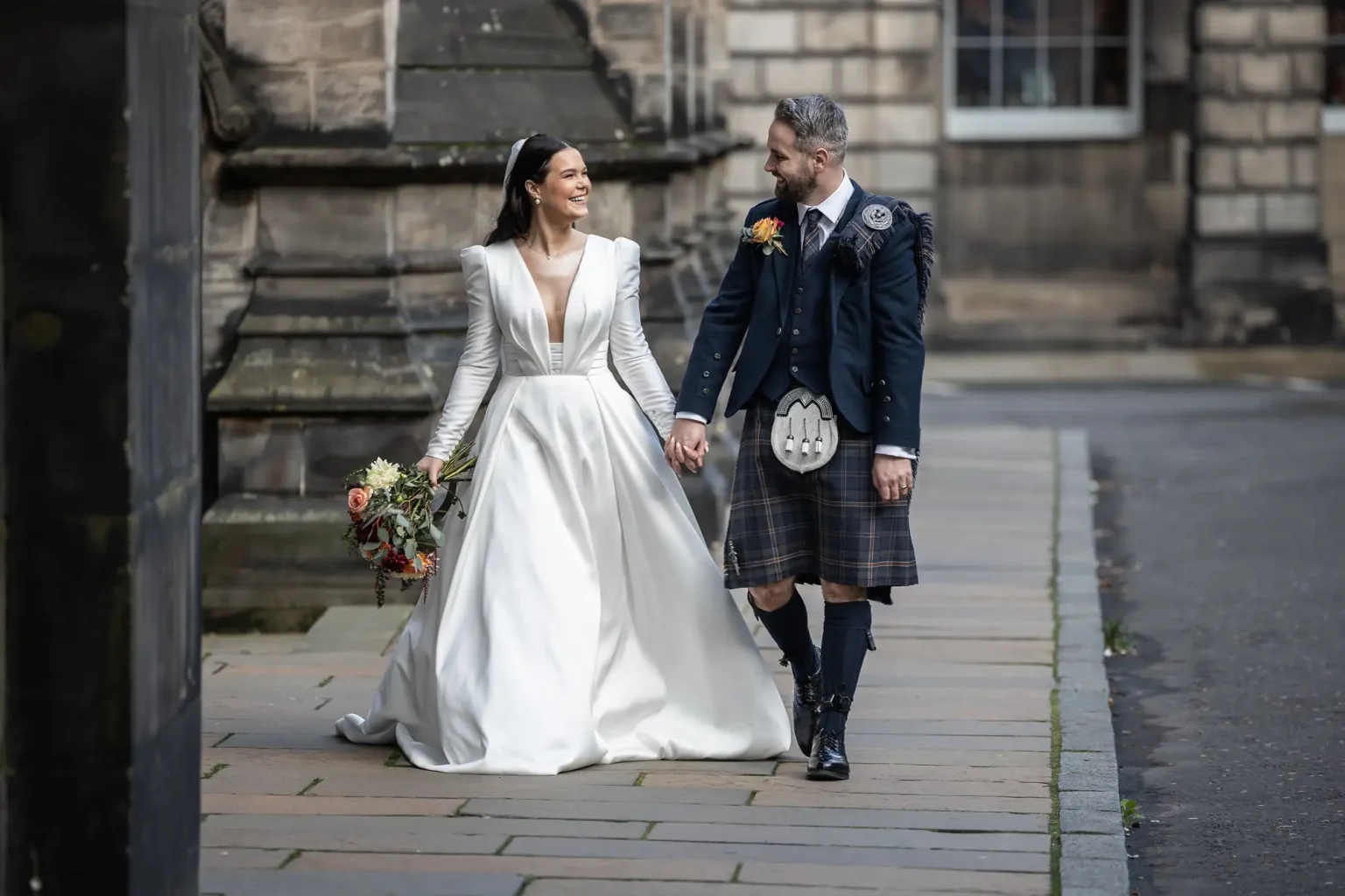 Natural photo of a couple at their wedding at The Signet Library, Edinburgh. A bride in a white gown and a groom in traditional Scottish attire hold hands and walk on a cobblestone path in a historic setting.