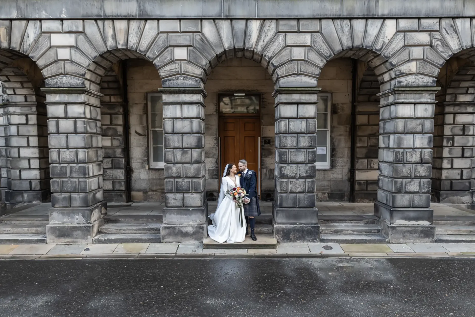 Newlywed couple posing in front of an ornate stone building with large arches. the bride holds a bouquet, and they stand close together, smiling.