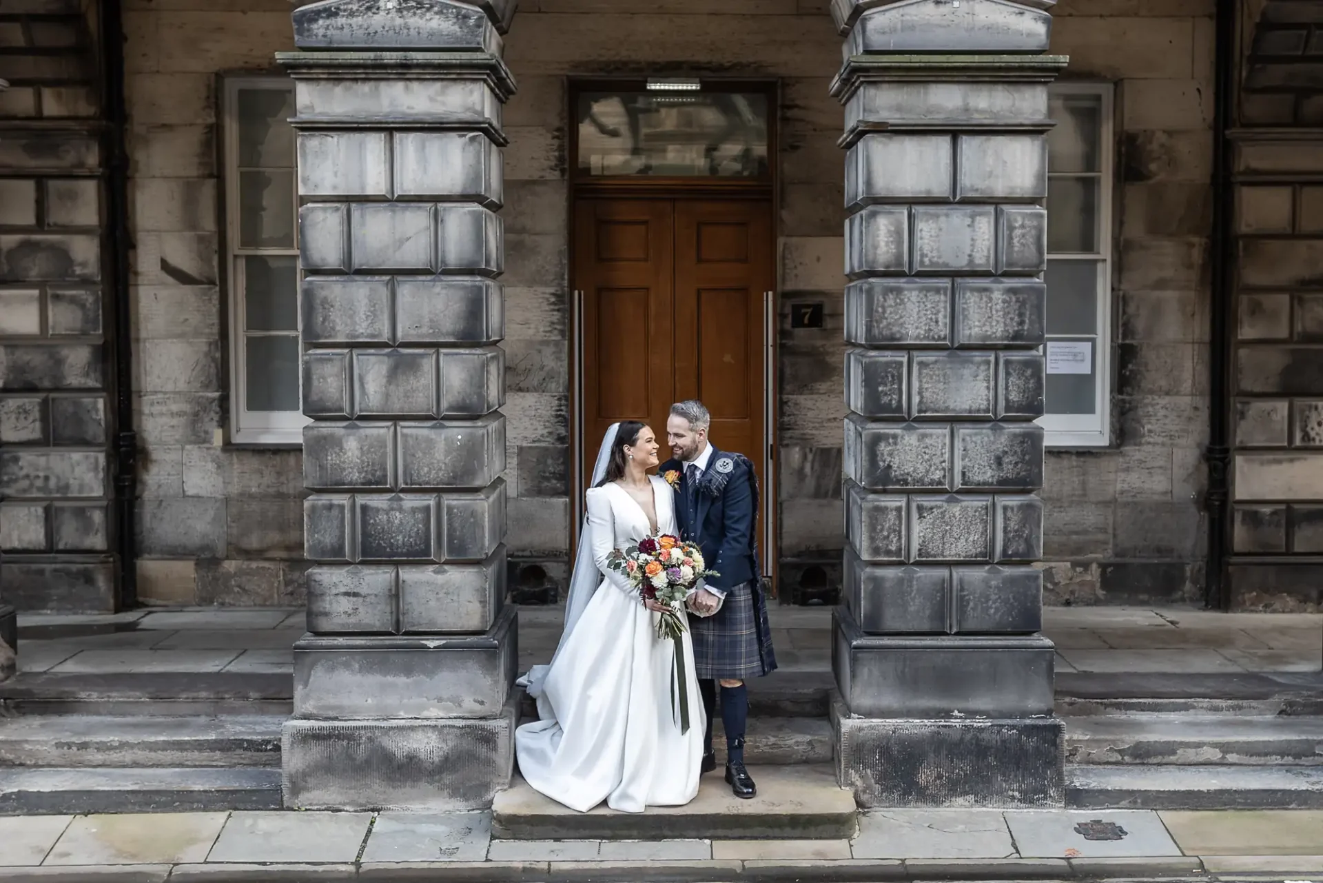 A bride and groom smiling and holding hands in front of a building with large stone columns and a wooden door.