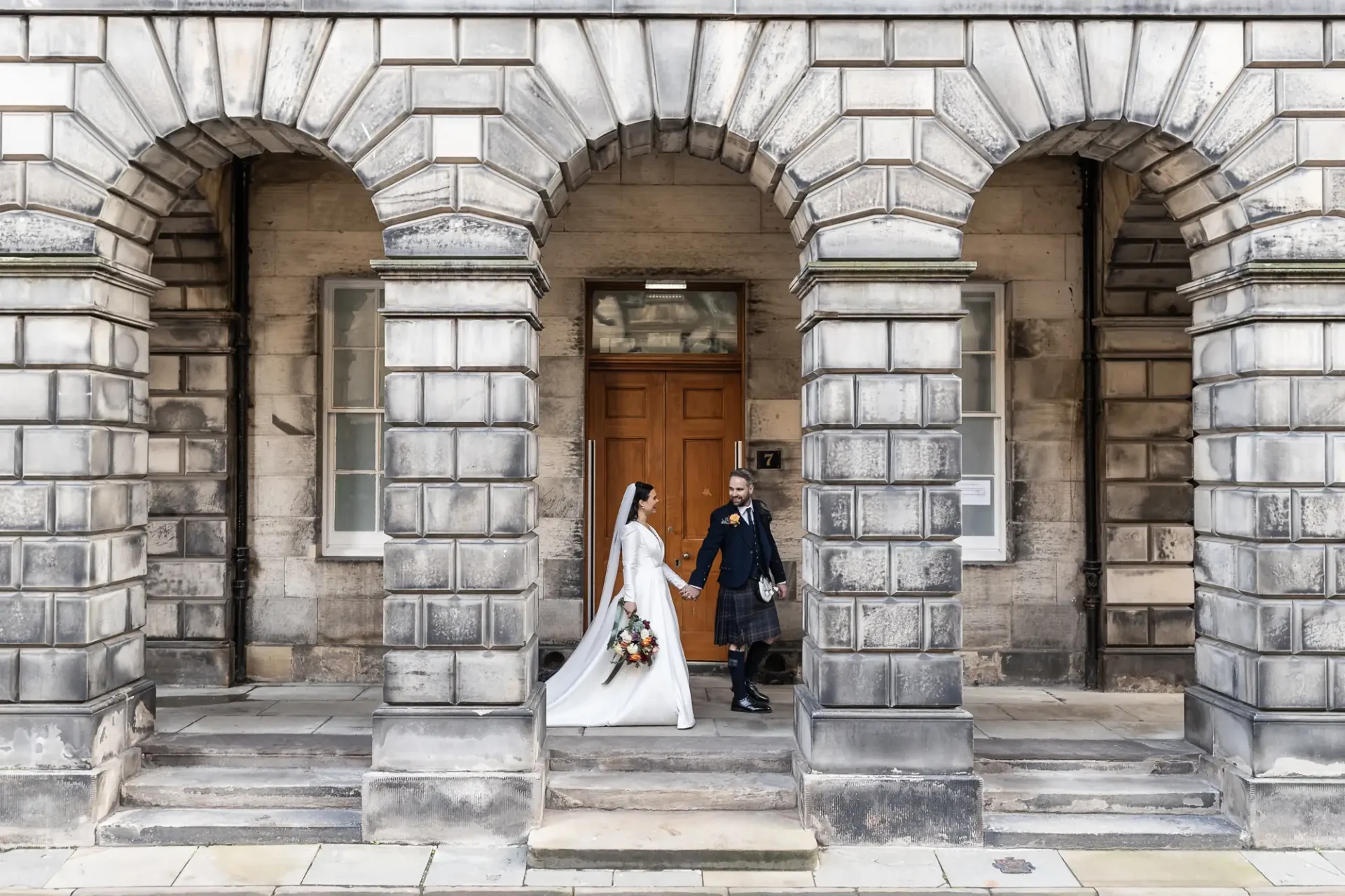 A bride and groom walking hand in hand through a grand stone archway, the groom in a military uniform and the bride in a white dress holding a bouquet.