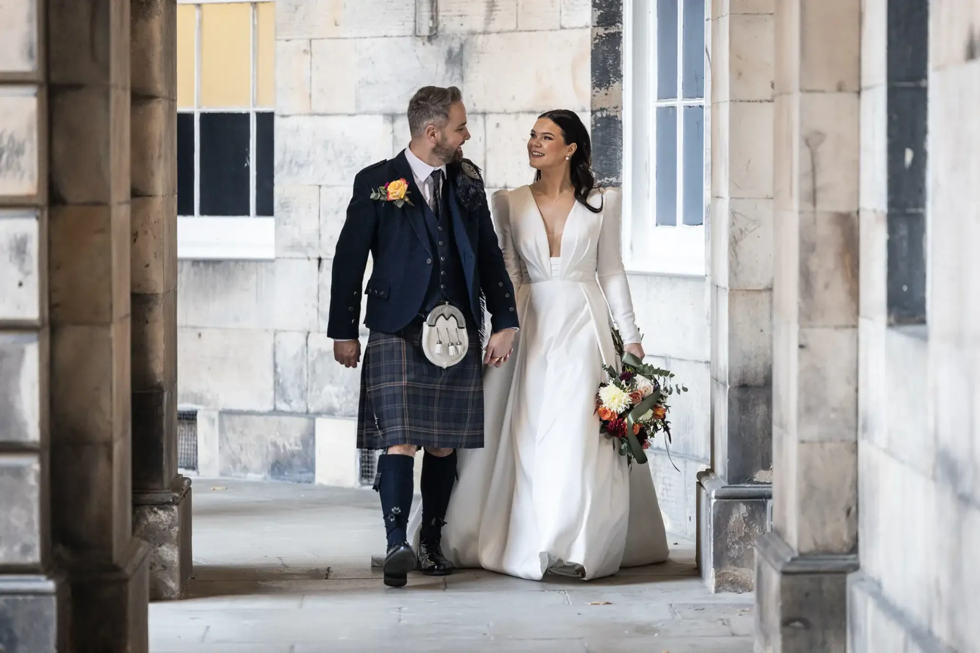A bride in a white dress and a groom in a kilt exchange smiles while walking hand in hand in a stone corridor.