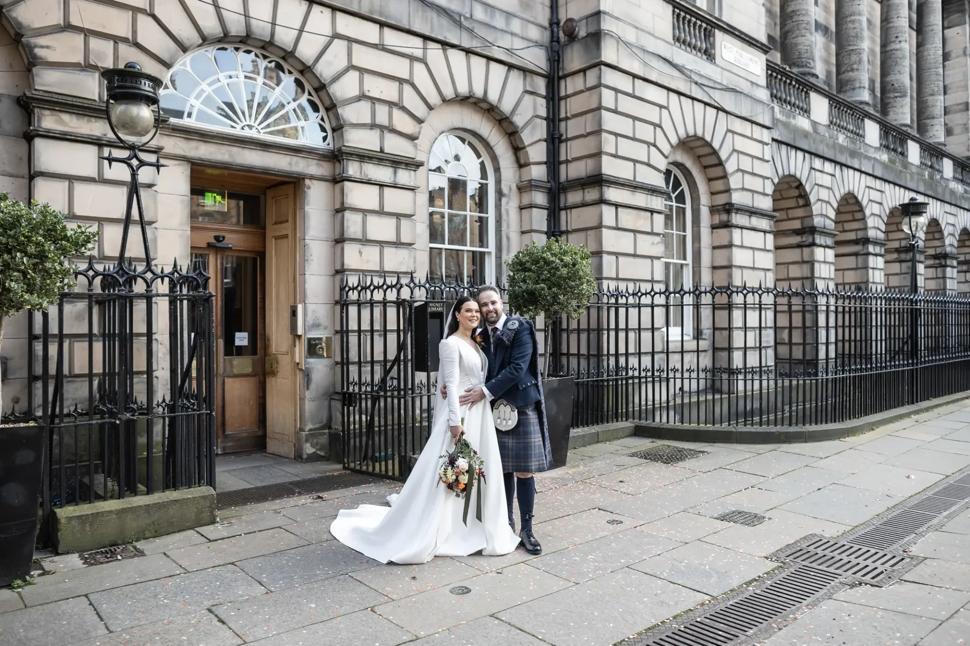 A bride and groom, the bride in a white dress and holding a bouquet, the groom in a kilt, smiling outside a grand stone building with columns and iron fences.