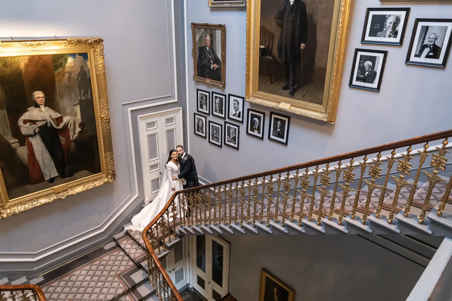 A couple poses on a grand staircase surrounded by numerous framed portraits hanging on the walls in an elegant building.