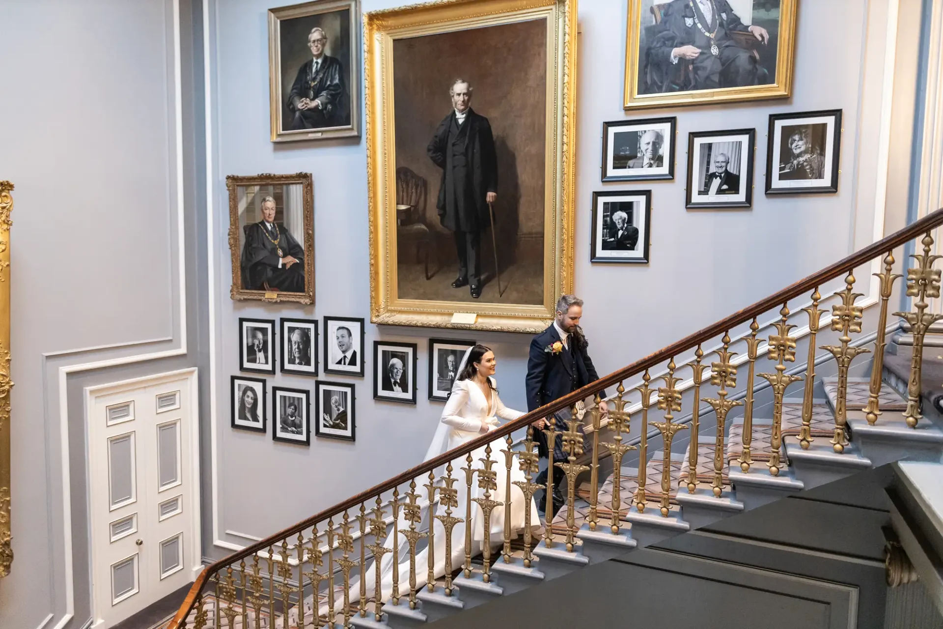 Two people ascending a grand staircase adorned with numerous framed portraits on the walls in a sophisticated gallery setting.