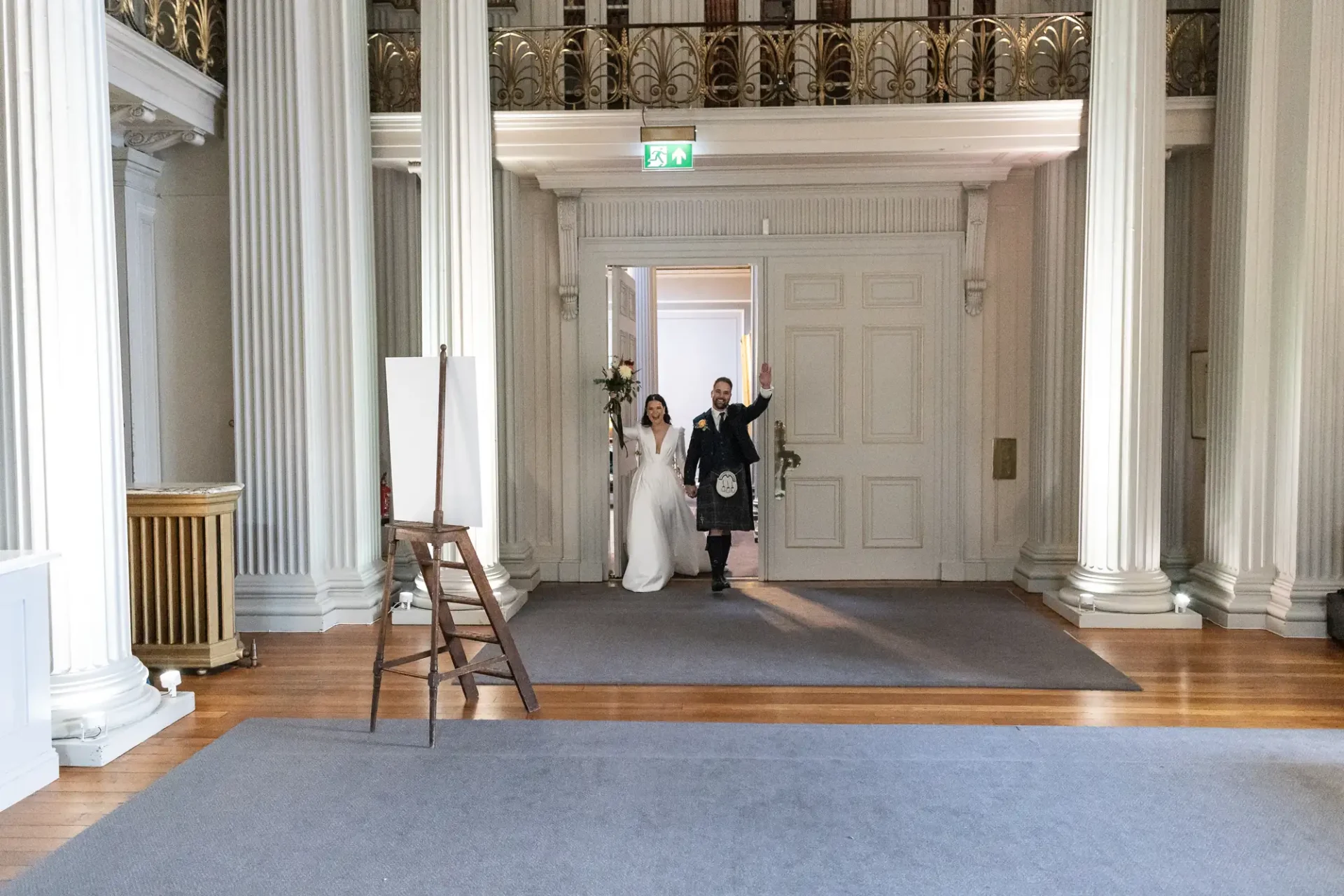 A bride in a white dress and a groom in a kilt walk joyfully through a grand hall doorway, holding hands.