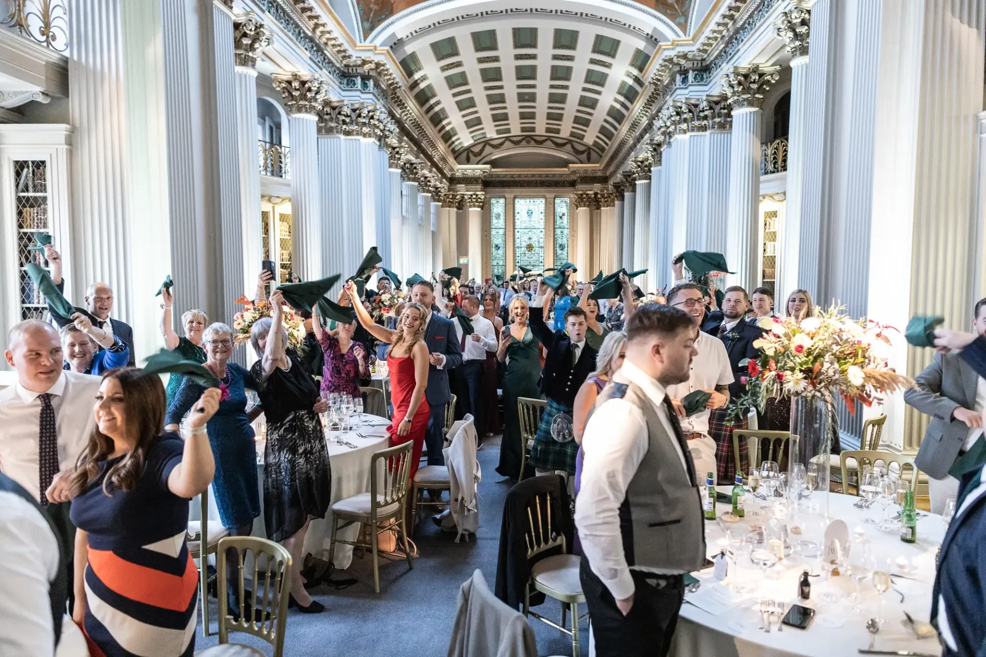 Guests waving napkins at a lively formal dinner in an elegant hall with classical columns and arched ceiling.