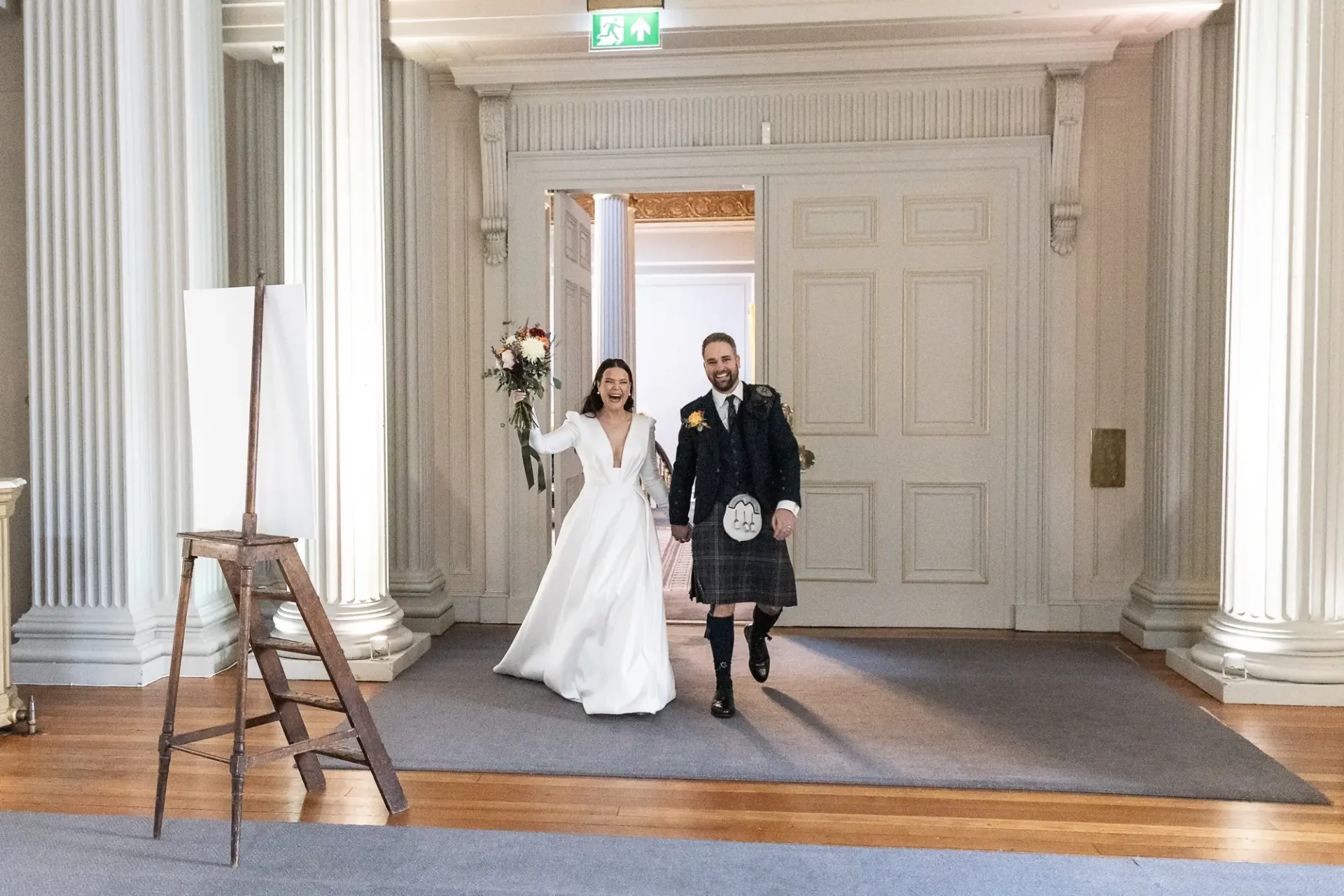 A bride in a white gown and a groom in a kilt holding hands and walking joyfully through an elegant room with classical architecture.