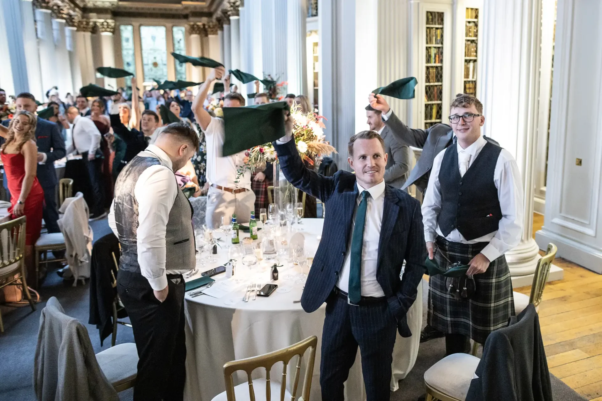 A man in formal attire standing and smiling at a lively event, holding a bouquet, as other attendees in kilts look on in an elegant dining room.