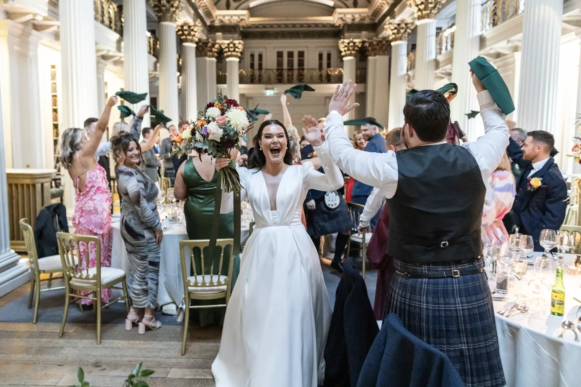 Bride and groom joyfully entering a grand dining hall, greeted with applause by guests, bride in white, groom in kilt.