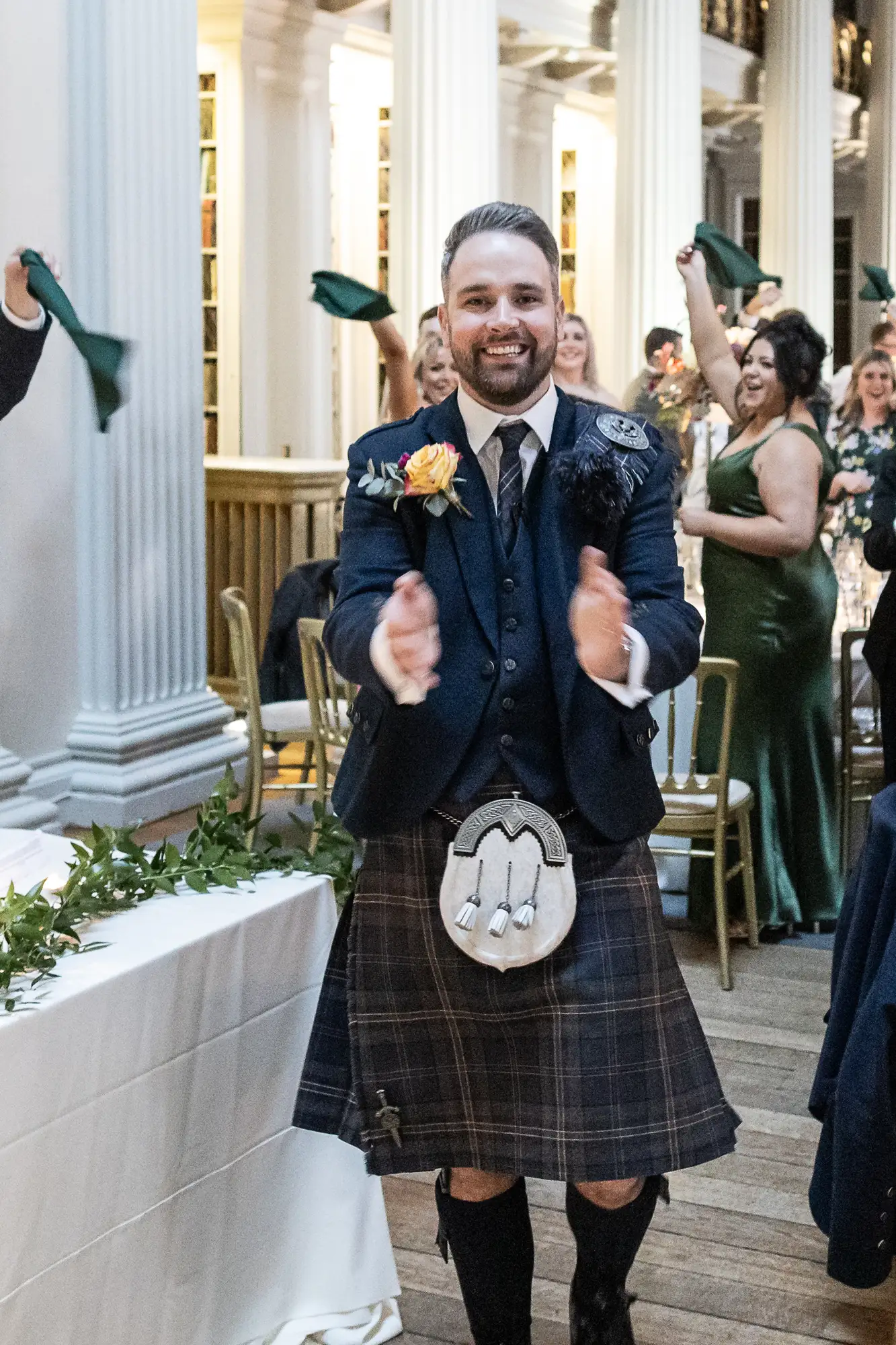 A smiling man in a traditional scottish outfit, including a kilt and sporran, walks enthusiastically down an aisle as guests applaud.