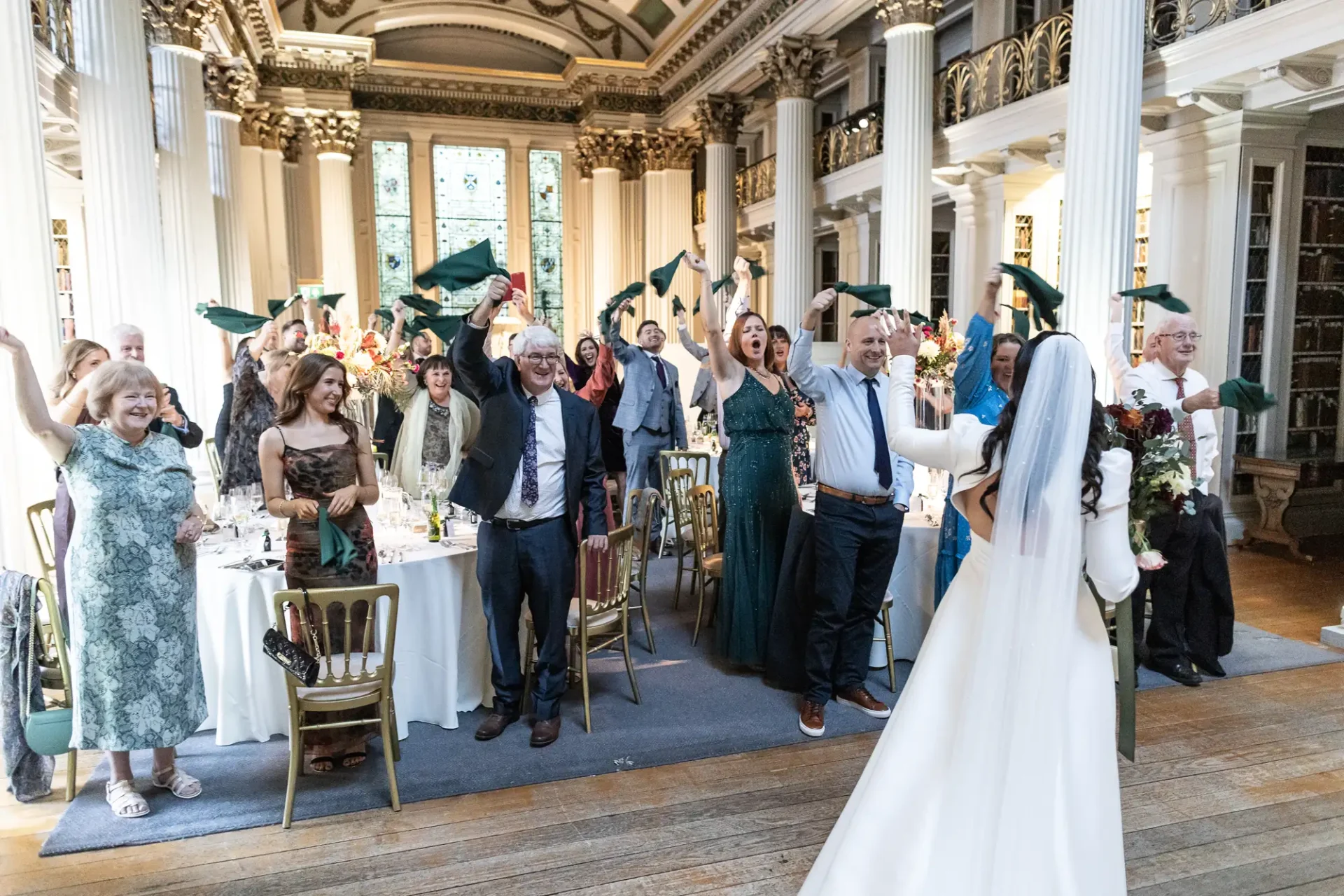 Wedding guests cheer and wave napkins as a happy couple walks down the aisle in a grand, ornately decorated hall.