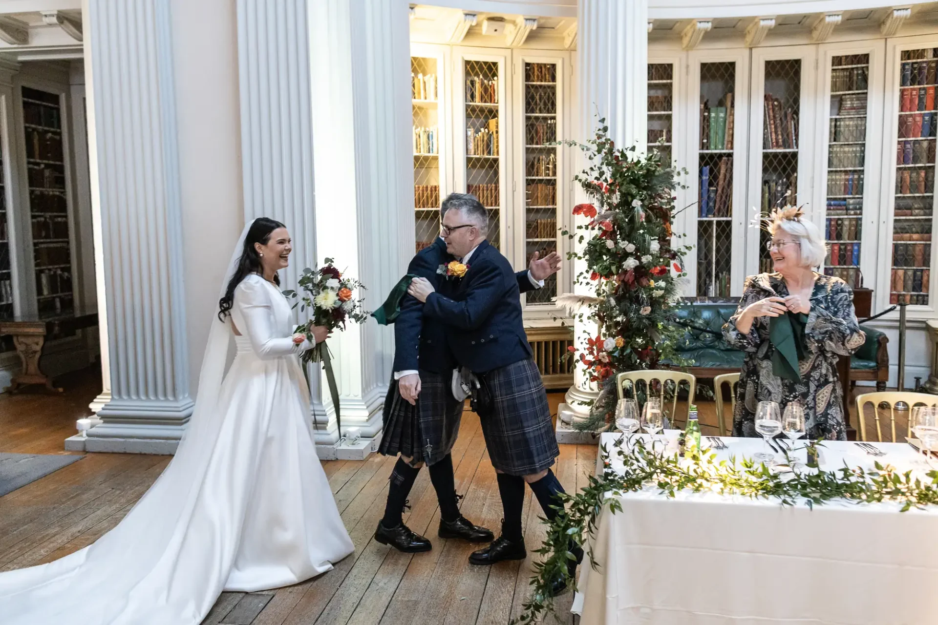 A bride in a white dress and a groom in a kilt laugh joyfully during a wedding toast in a library with guests applauding.