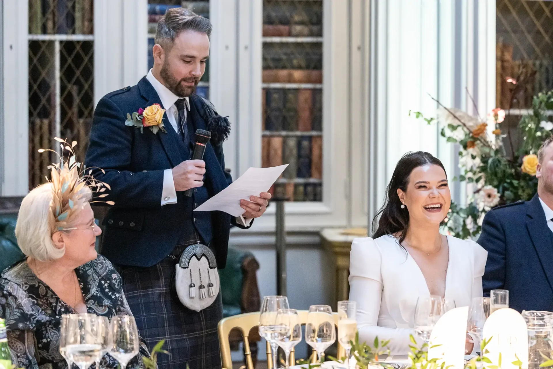 A man in a kilt delivers a speech at a wedding, holding a paper, as a bride sitting next to an older woman laughs joyfully at a decorated table.