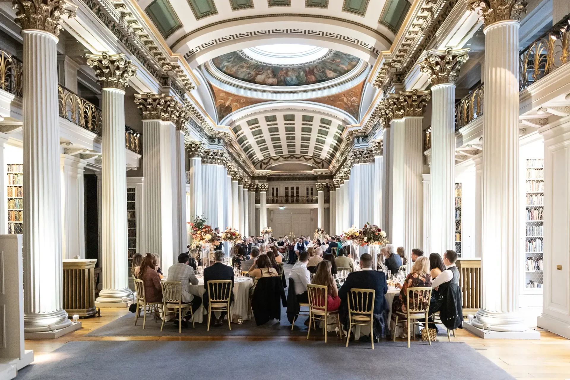 Guests seated at a formal event in an ornate hall with rows of columns and a detailed ceiling.