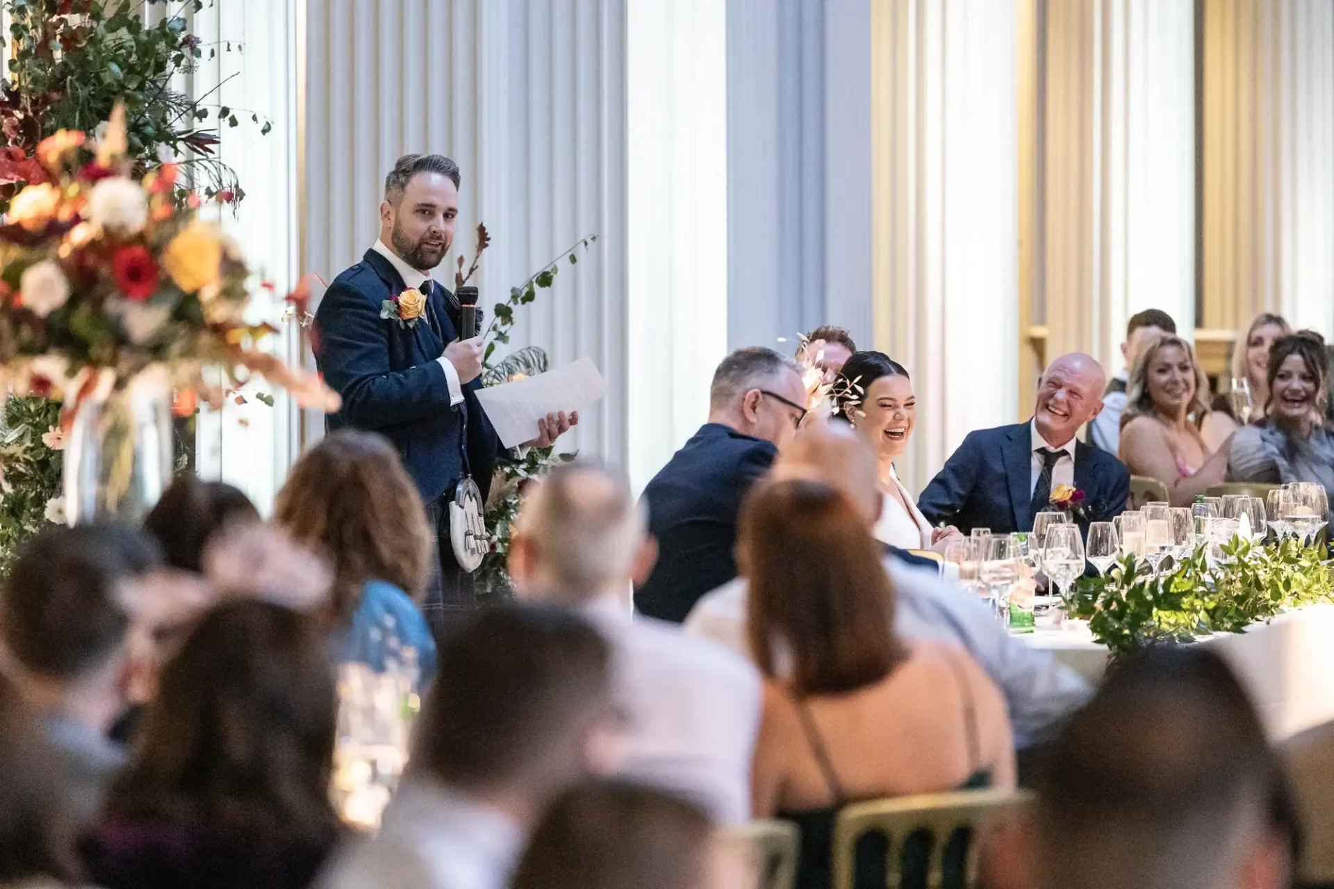 Man giving a speech at a wedding reception, holding a microphone, with guests seated at tables listening and smiling.