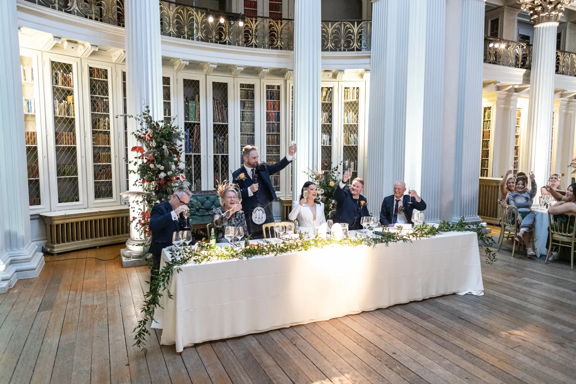 A wedding reception in an elegant hall with guests watching as a man standing gestures joyfully at the head table.