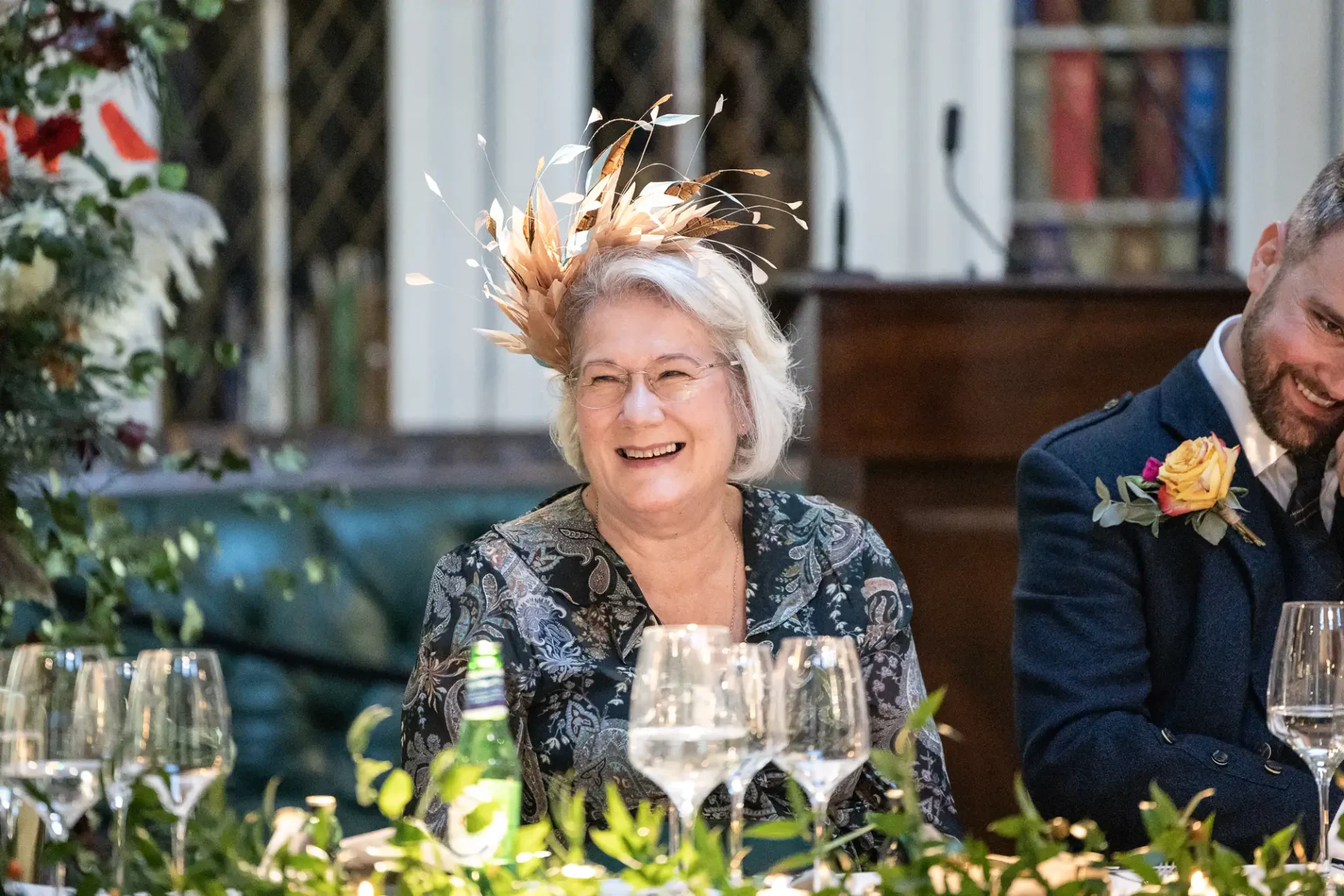 An elderly woman in a floral dress and feathered hat laughs joyfully at a wedding reception table, beside a smiling man in a suit with a boutonniere.