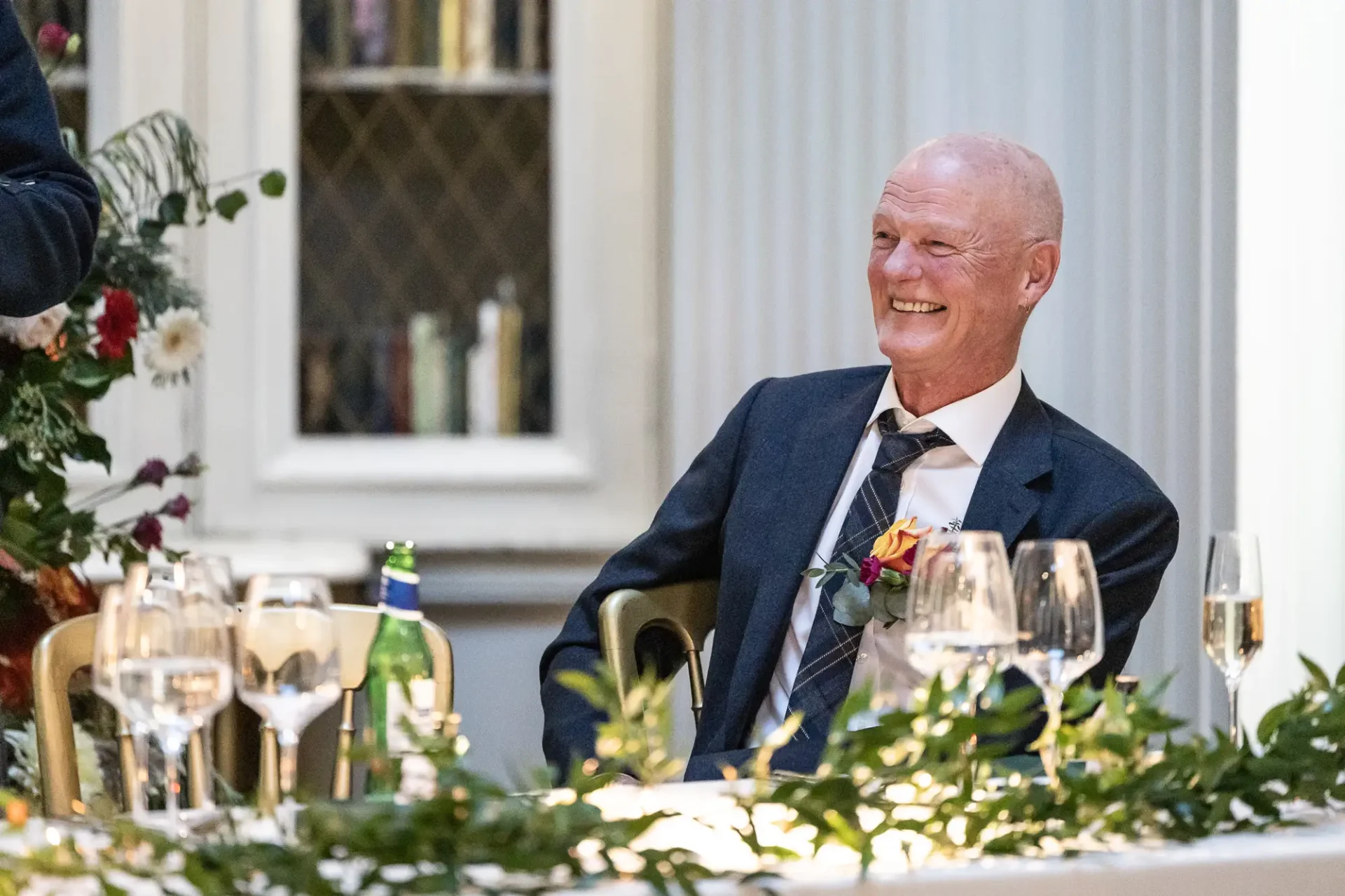 An elderly man in a suit, laughing at a formal dinner table adorned with flowers and wine glasses.