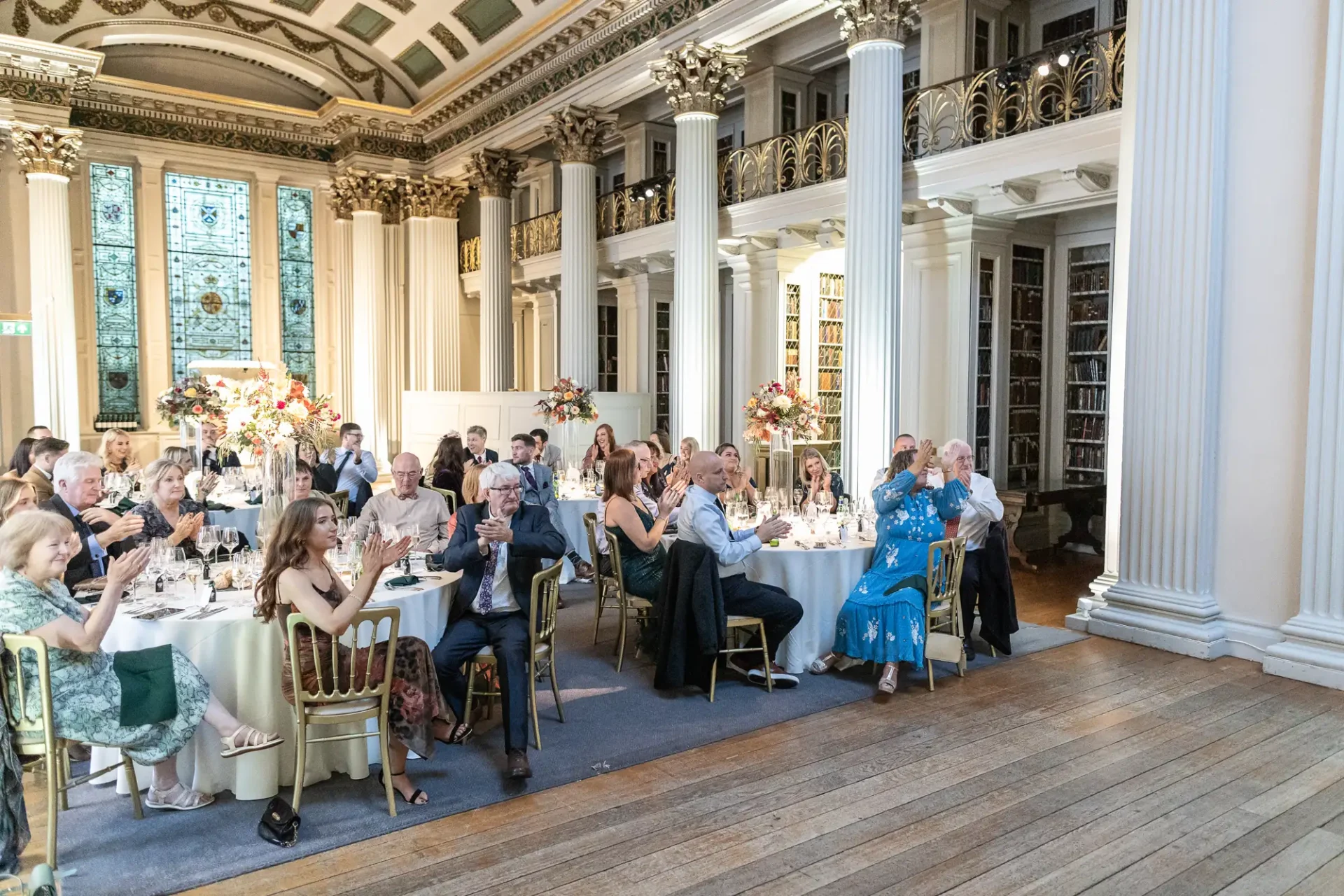 Guests seated at banquet tables applauding in an elegant hall with ornate balconies and a stained glass window.