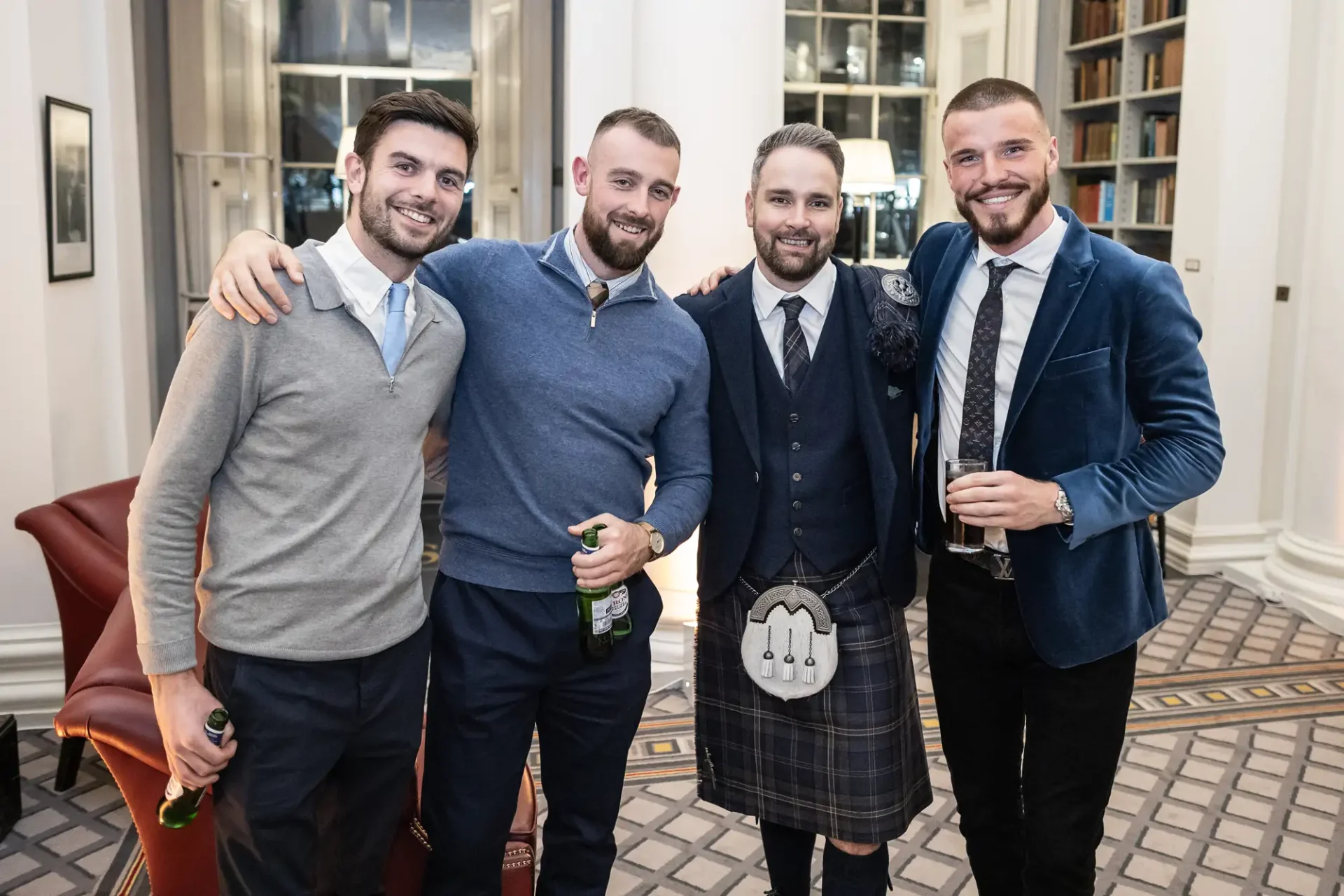 Four men smiling at a formal event, one wearing a kilt, holding drinks in a library setting with elegant decor.
