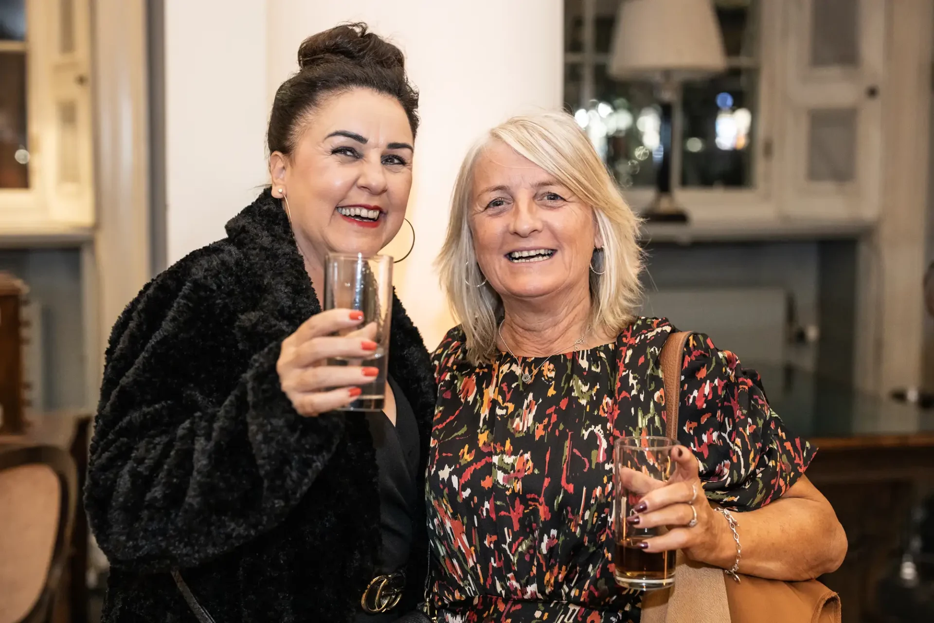 Two women smiling and holding champagne glasses at a social event indoors. one is wearing a black fur coat, and the other a floral dress.