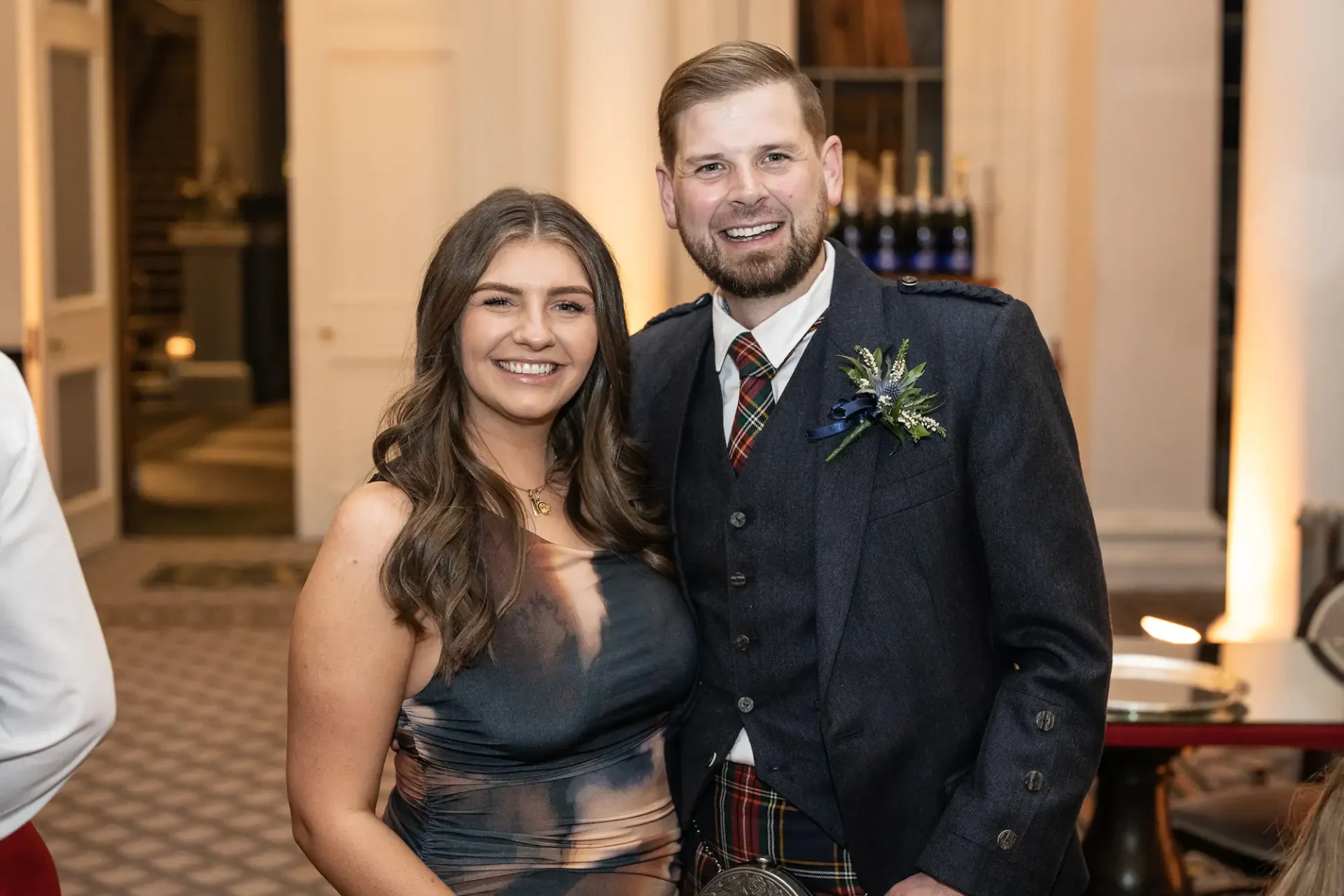 A man in a tartan kilt and jacket with a woman in a gray dress, both smiling, standing together at an indoor event.