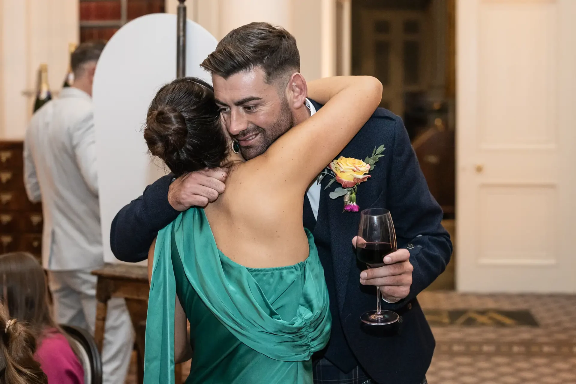 Man in a suit hugs a woman in a green dress at a formal event, holding a glass of red wine.