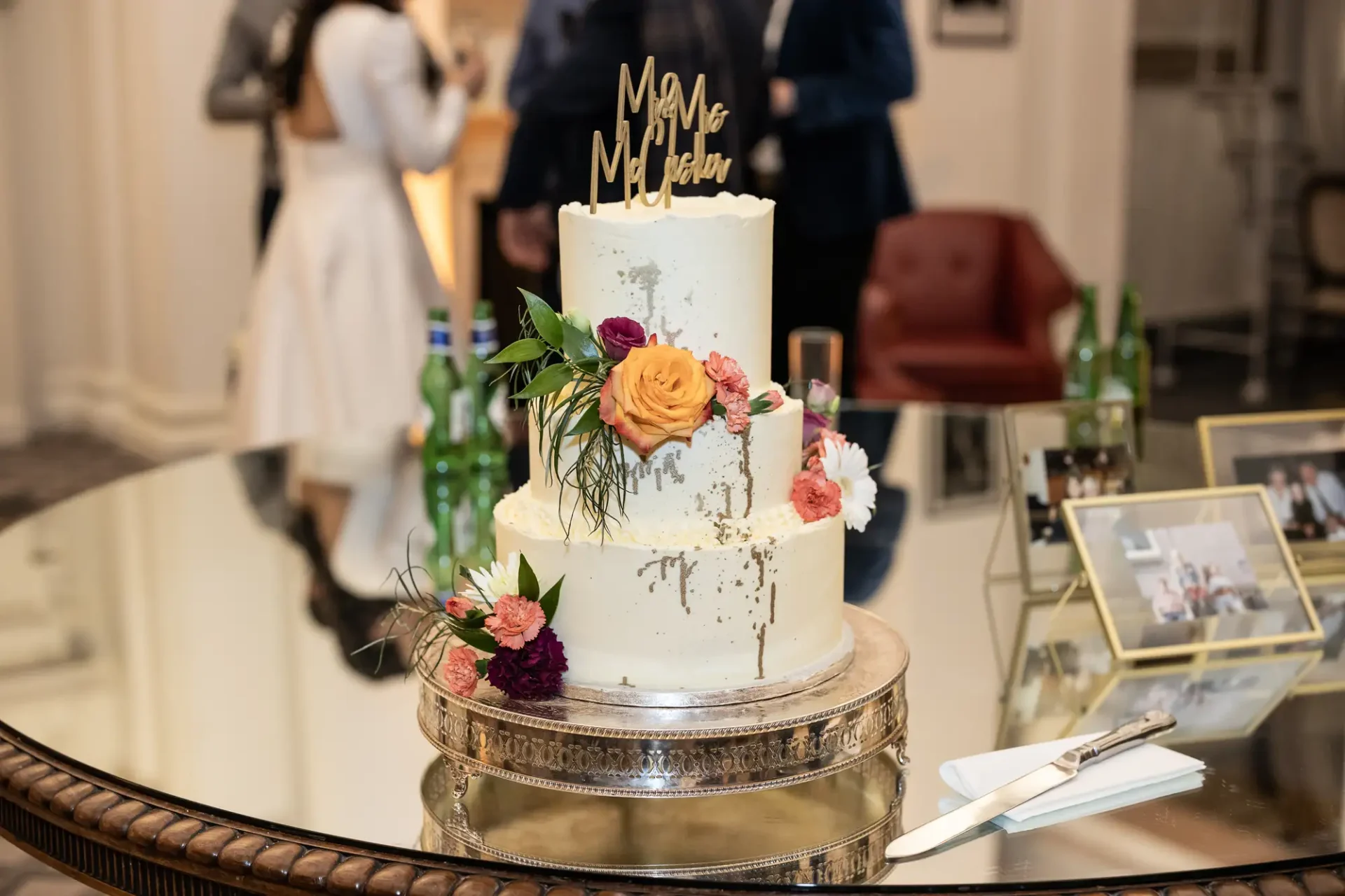 A two-tiered wedding cake decorated with fresh flowers on a glass table, with a "mr & mrs" topper, surrounded by guests in elegant attire at a reception.