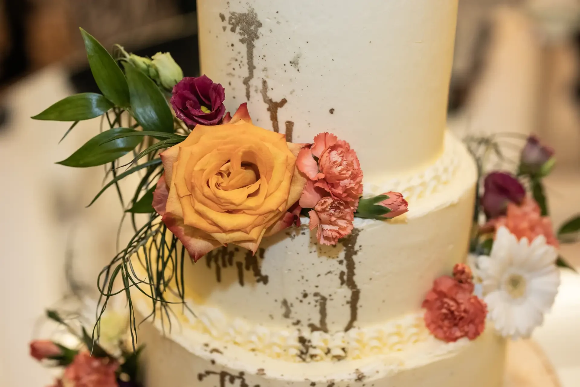 Close-up of a three-tiered white wedding cake adorned with fresh flowers, including an orange rose and pink carnations, with light frosting drips.