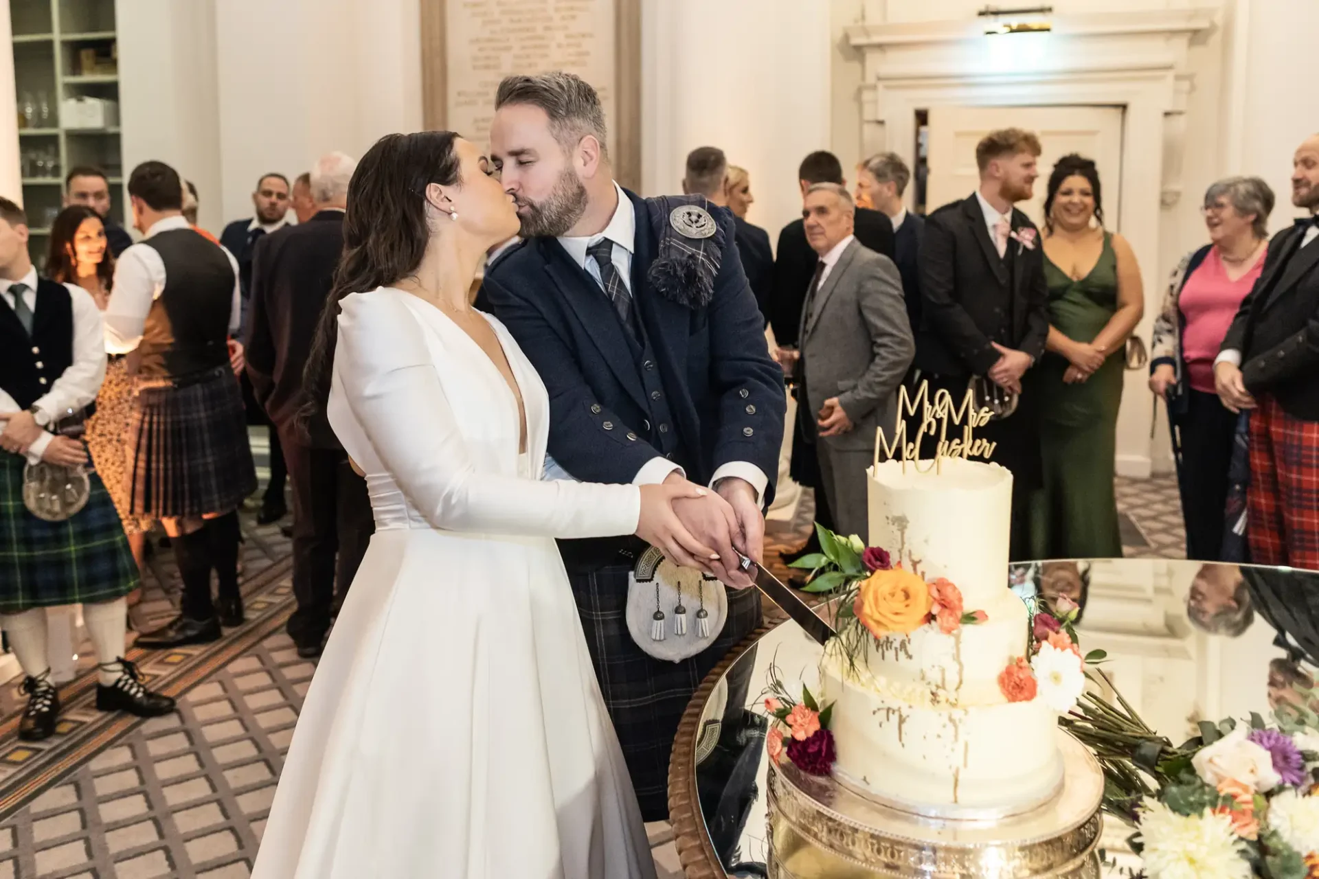 A bride and groom in traditional scottish attire kiss while cutting a wedding cake, surrounded by guests in a decorated hall.