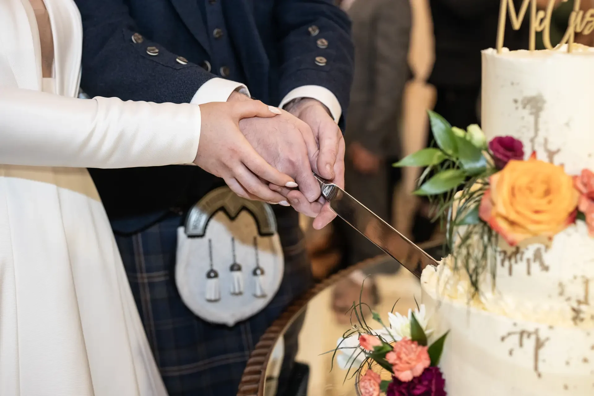 A couple in wedding attire cutting a cake together, with the person on the left in a white dress and the person on the right wearing a kilt.