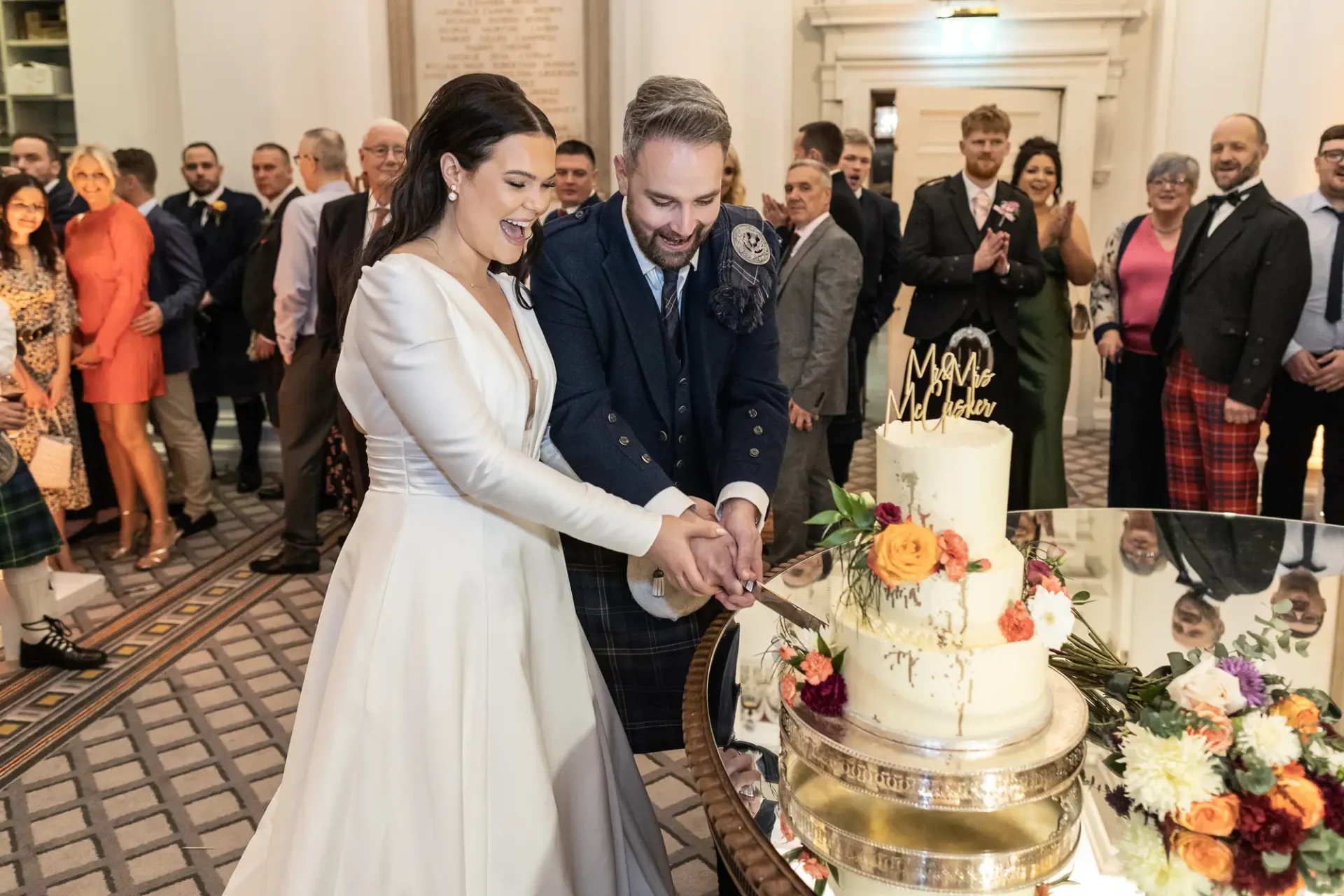 A bride and groom joyfully cutting a large, three-tiered wedding cake surrounded by guests in a decorated venue.