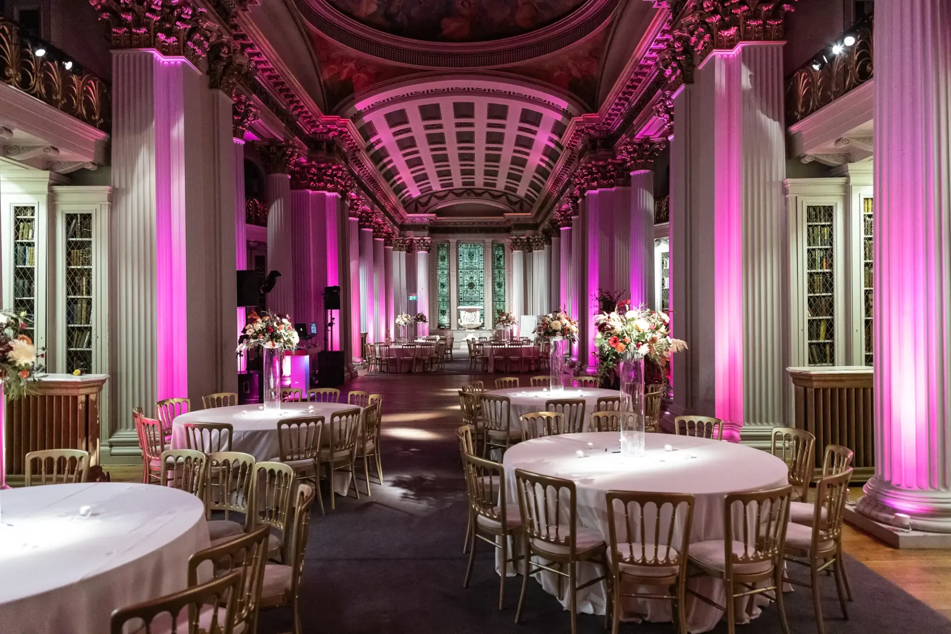 Elegant event hall with pink lighting, white round tables, and floral centerpieces, featuring grand arches and detailed ceiling.