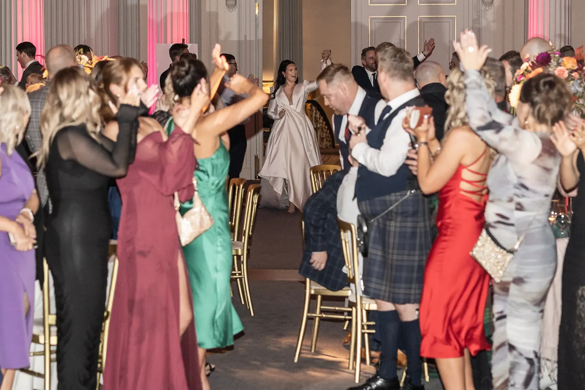 A bride and groom walking through a cheering crowd at a wedding reception, with guests in formal attire clapping and some wearing kilts.