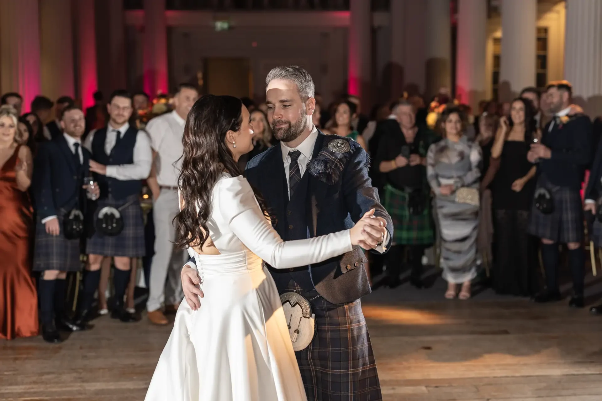 A couple in wedding attire dances in front of an audience, the man wearing a kilt and the woman in a white gown.